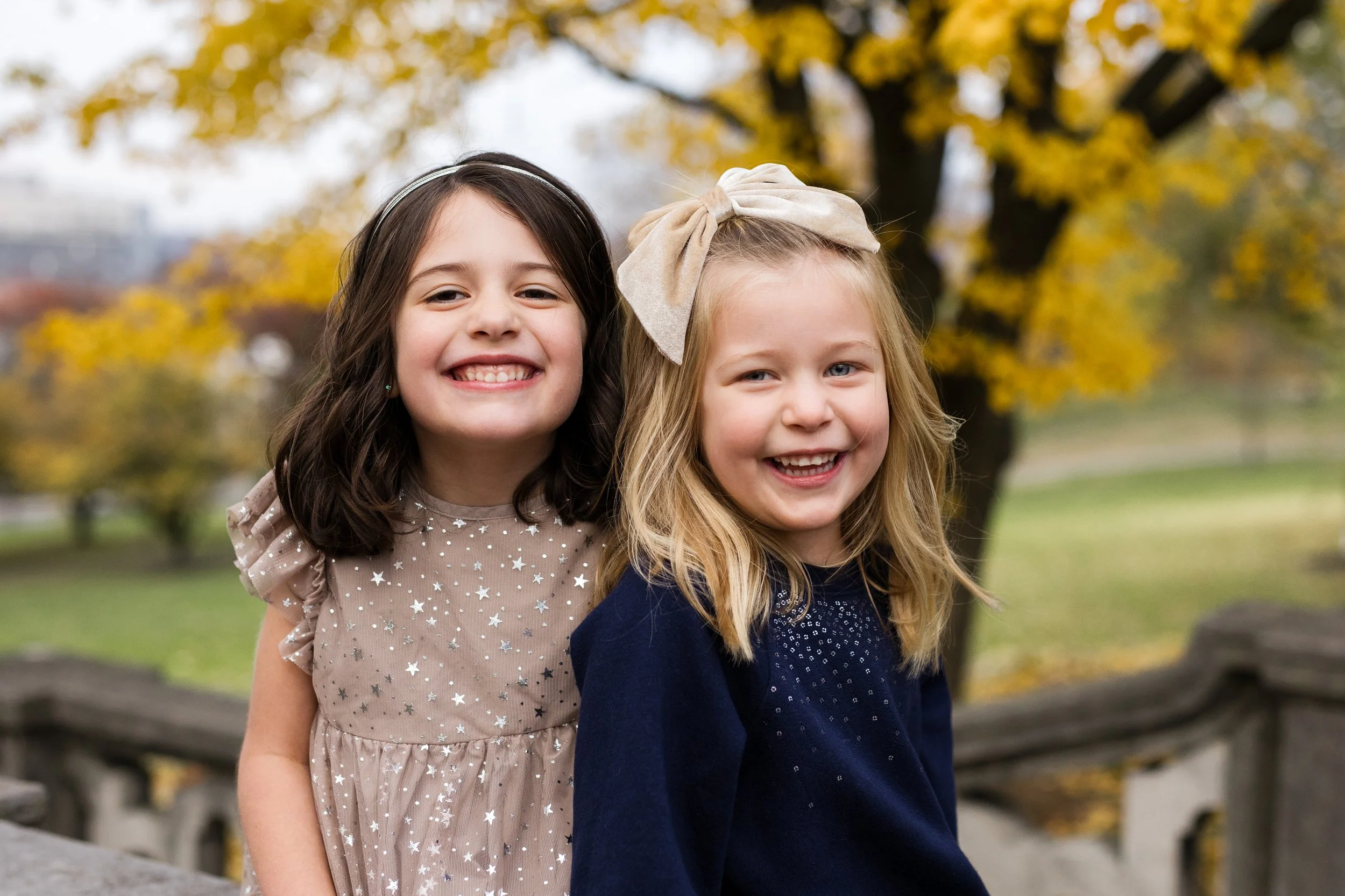2 sisters sit below a tree full of yellow fall leaves. They both have big smiles on their faces.