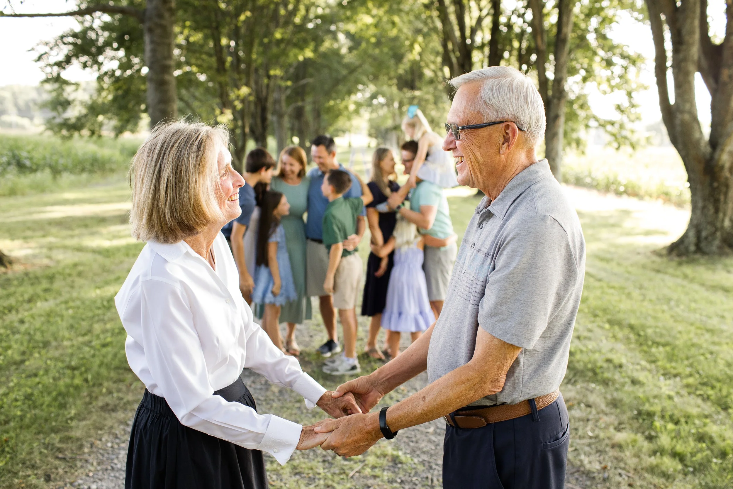 Grandparents hold hands and look at each other. In the distance, you see their kids and grandkids chatting together, happily.