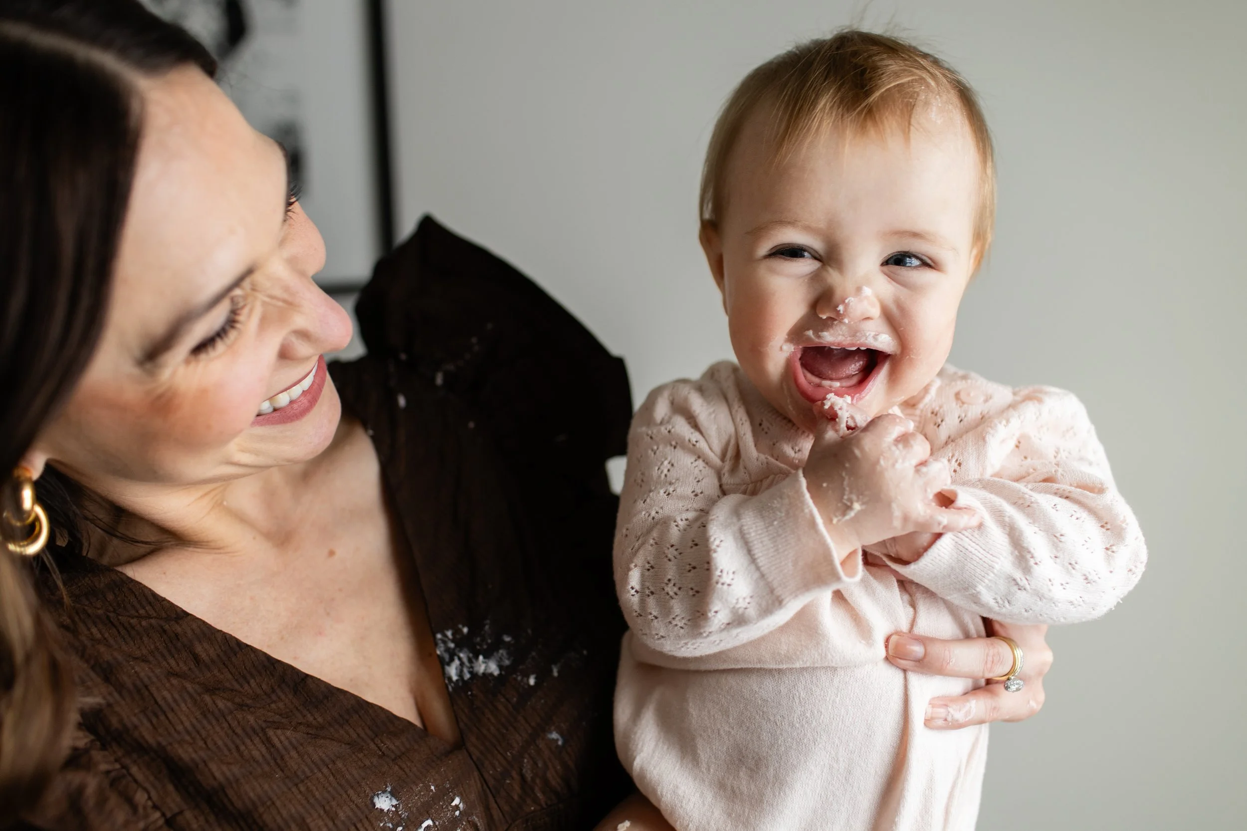 A mom holds her newly one-year-old. Both are covered in icing from a smash cake the baby devoured.