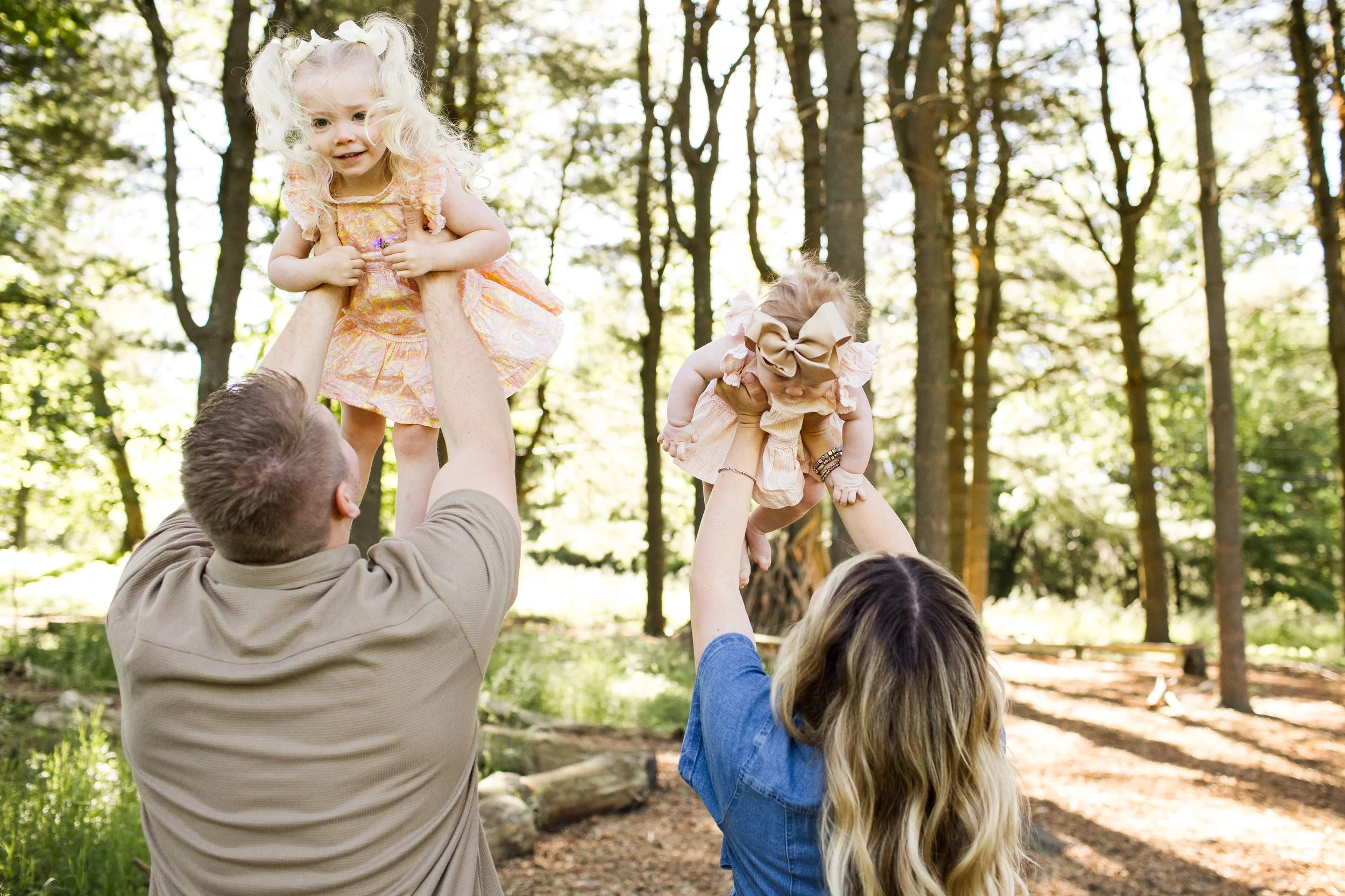 Parents lift up their 2 girls under a canopy of trees.