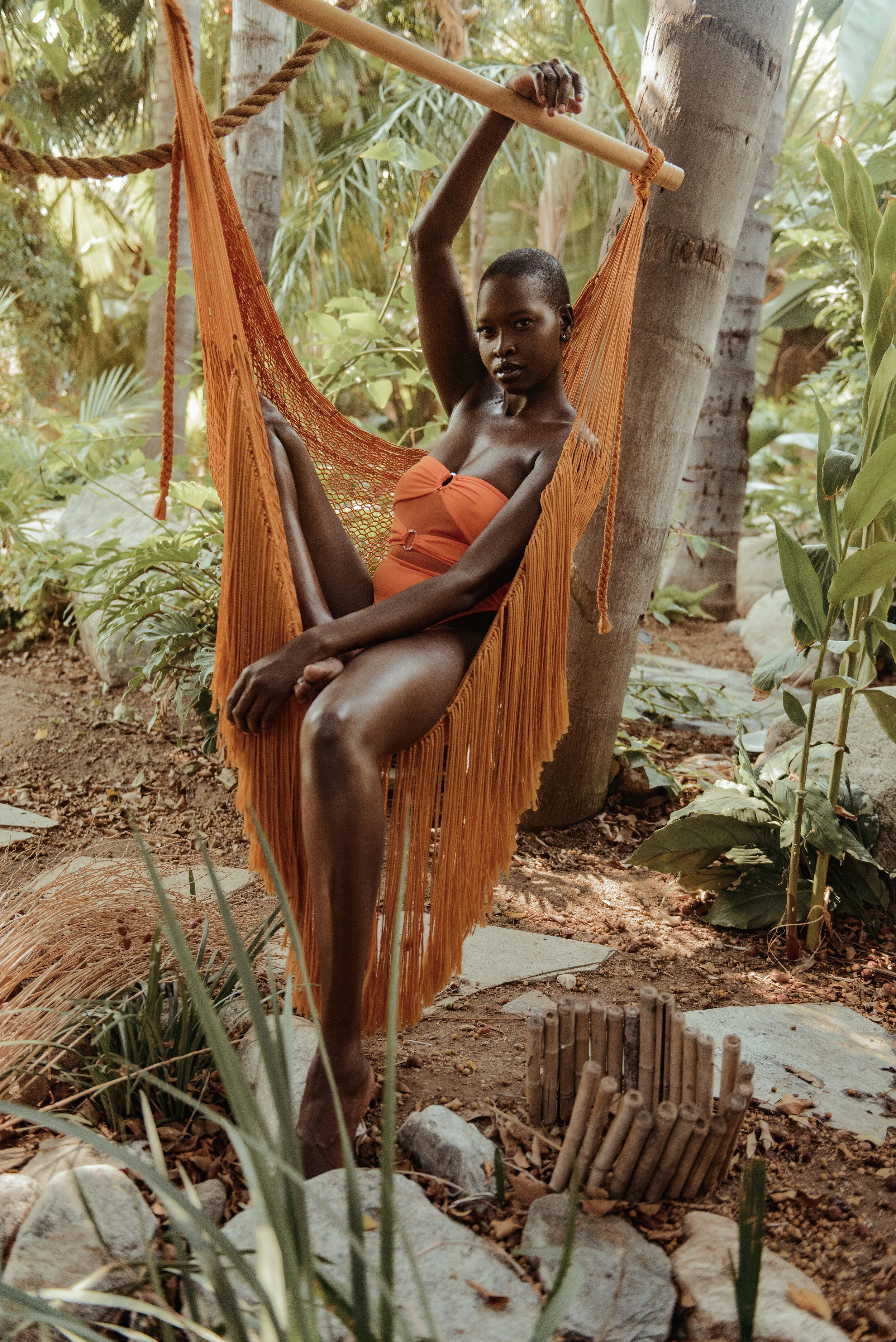  Tropical resortwear portrait of model in burnt orange one-piece on woven hammock surrounded by greenery   