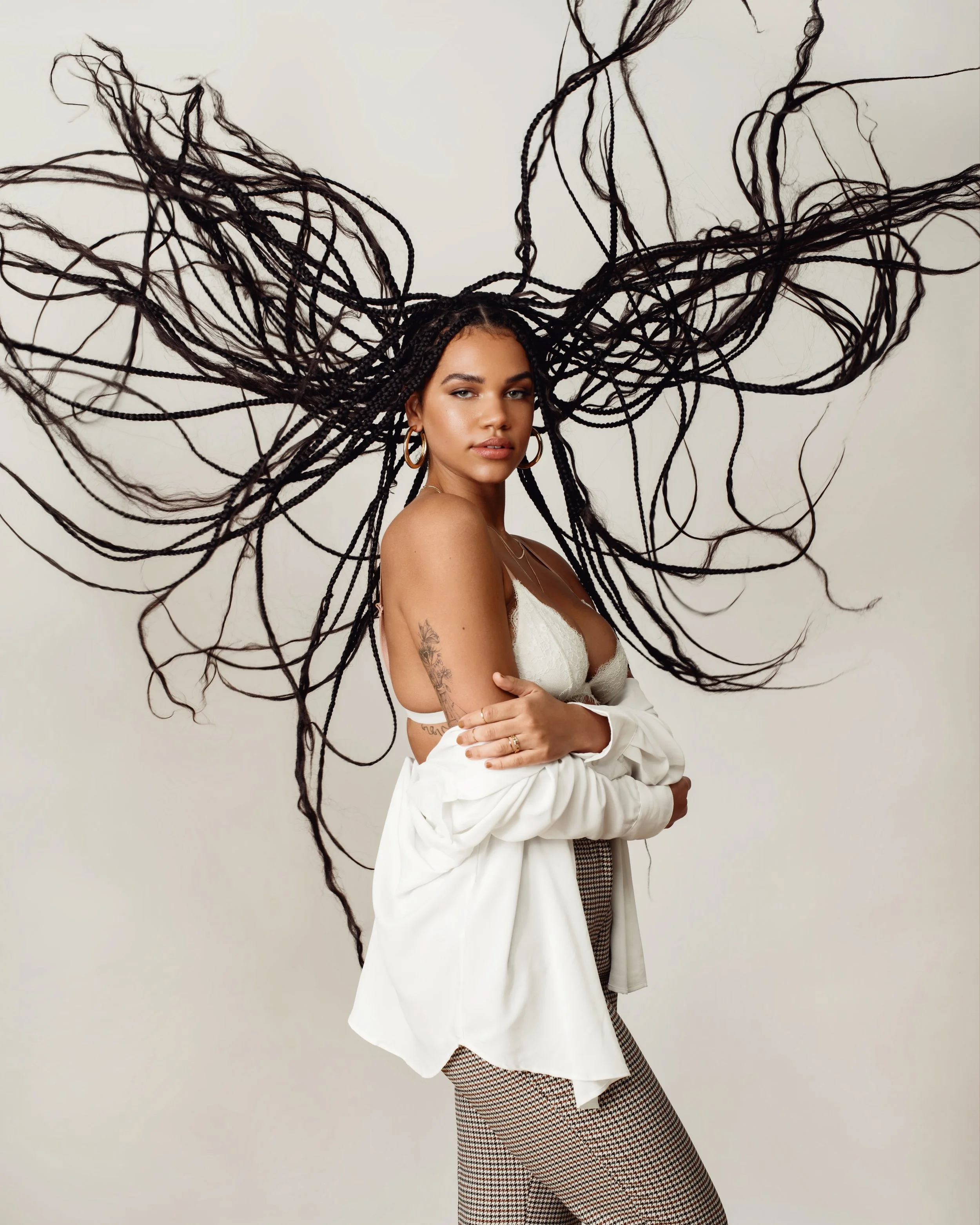 Studio editorial of model with long braids flying outward in motion while wearing white blouse and houndstooth leggings 