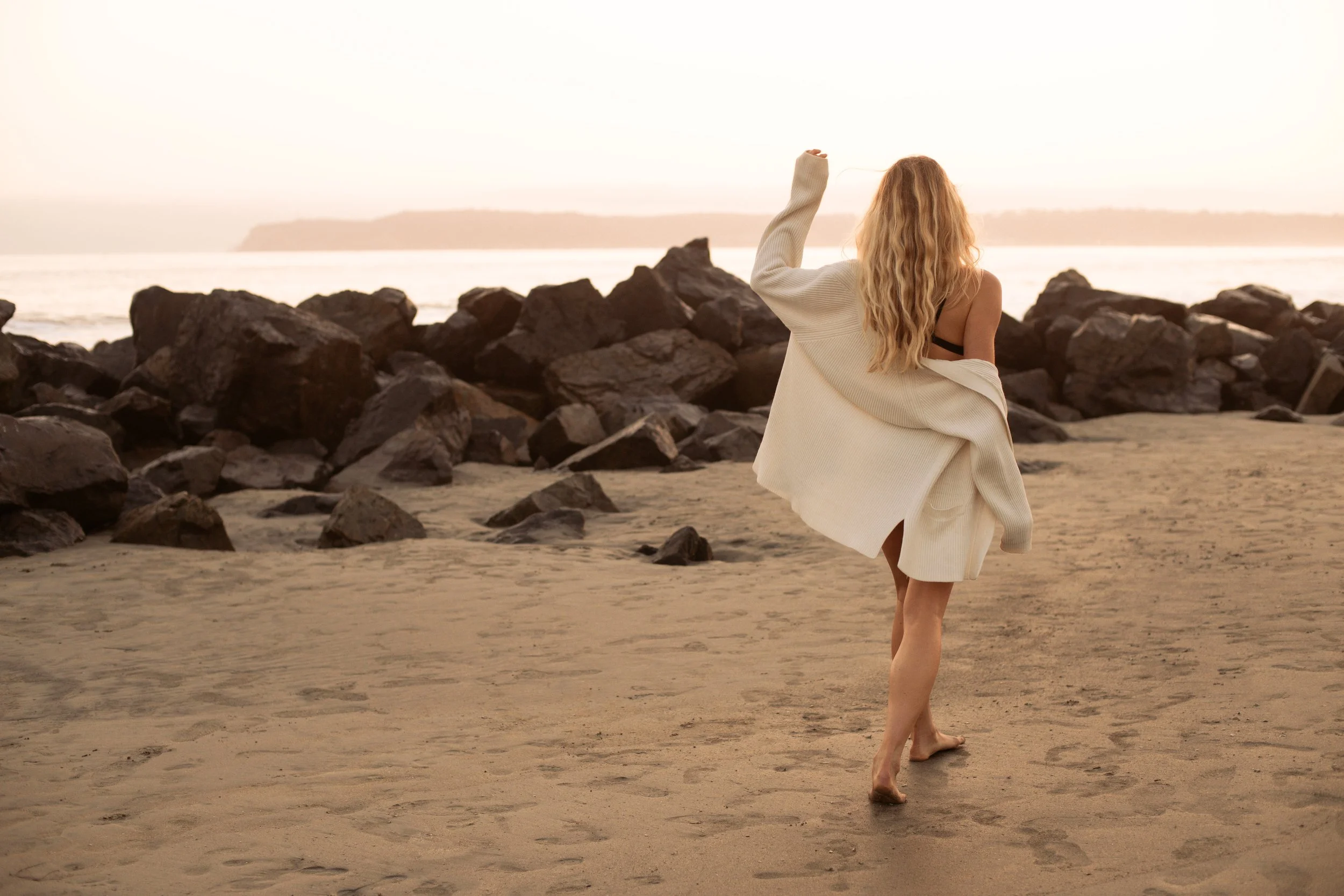  Golden hour lifestyle image of model in knit sweater and black swimwear walking across the sand 