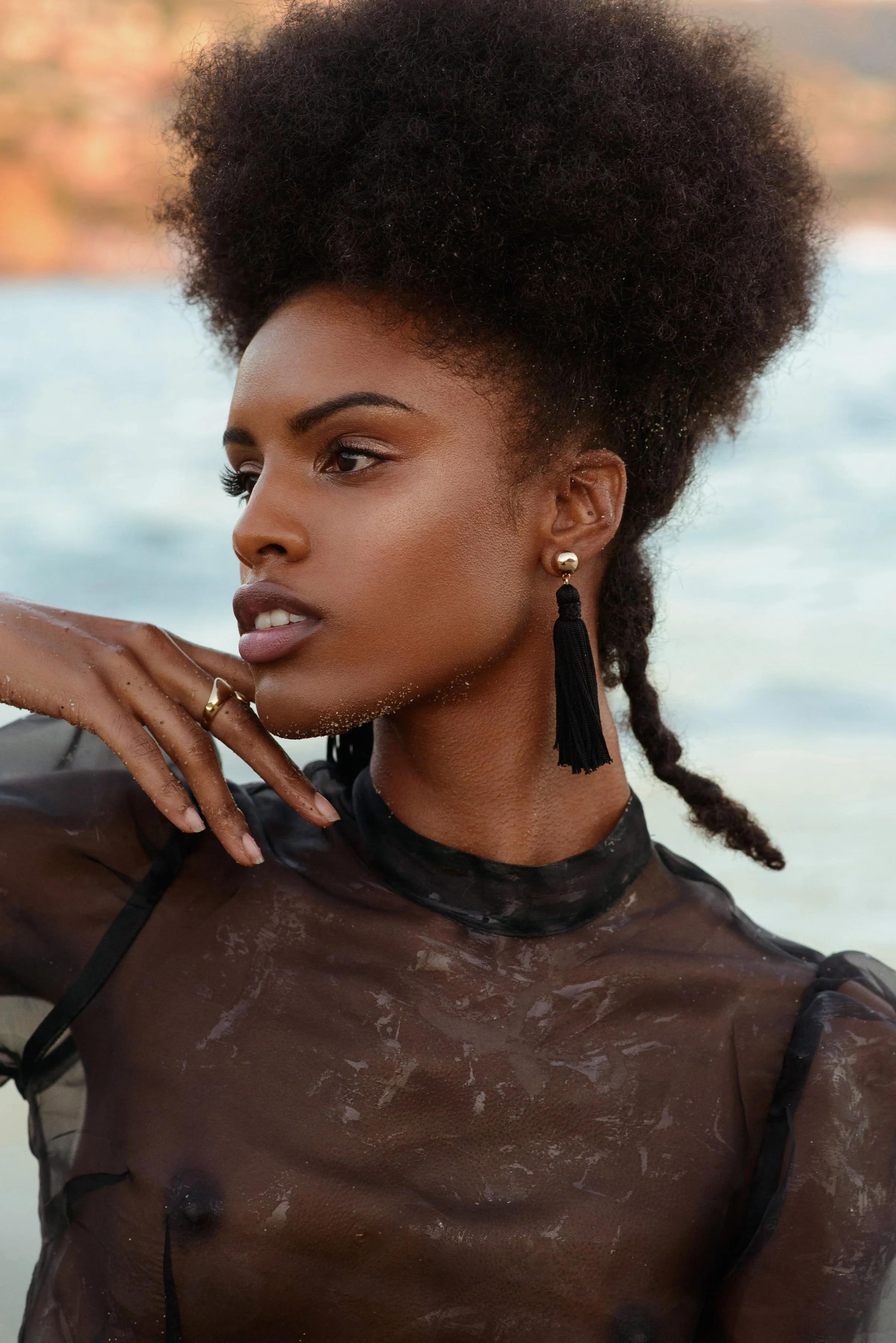  Close-up beauty editorial with model in sheer black top and statement tassel earrings by water 