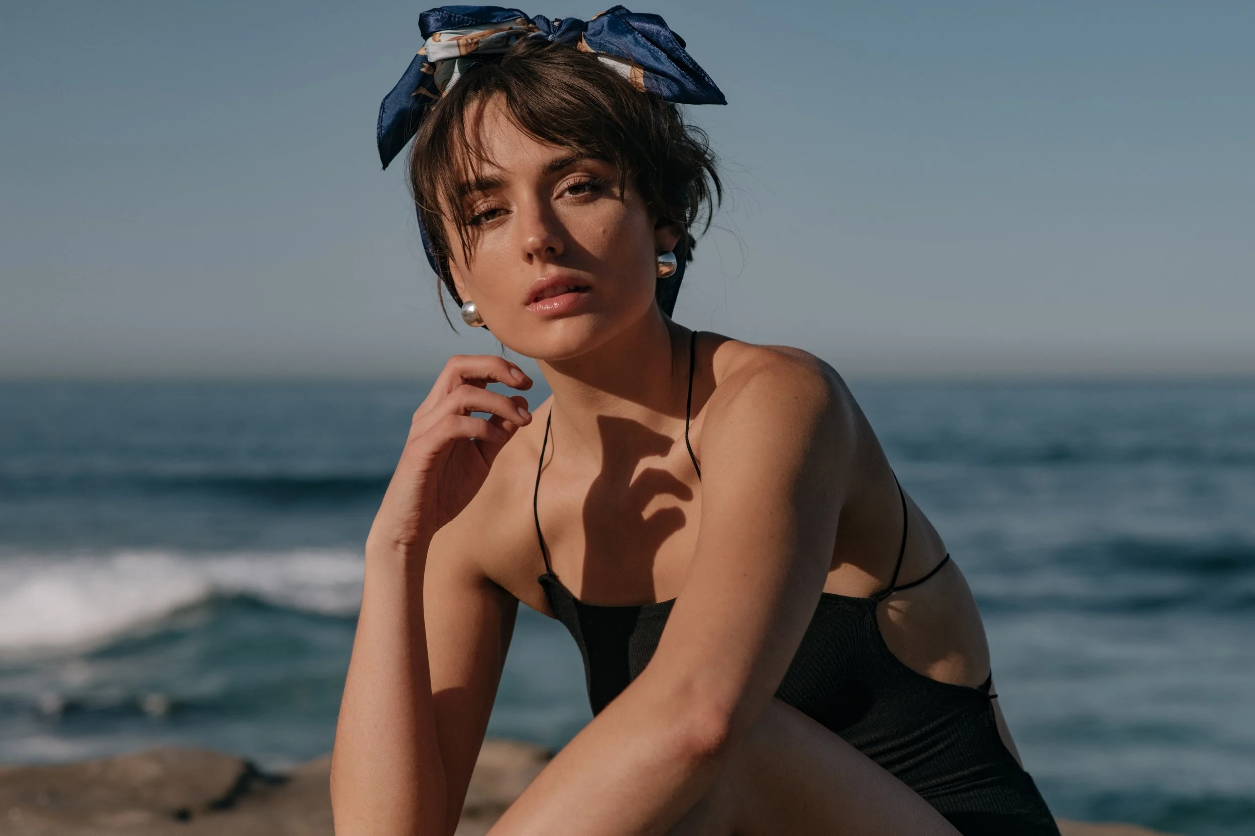  Close-up portrait of model in black cutout swimsuit and blue headscarf sitting by the sea 