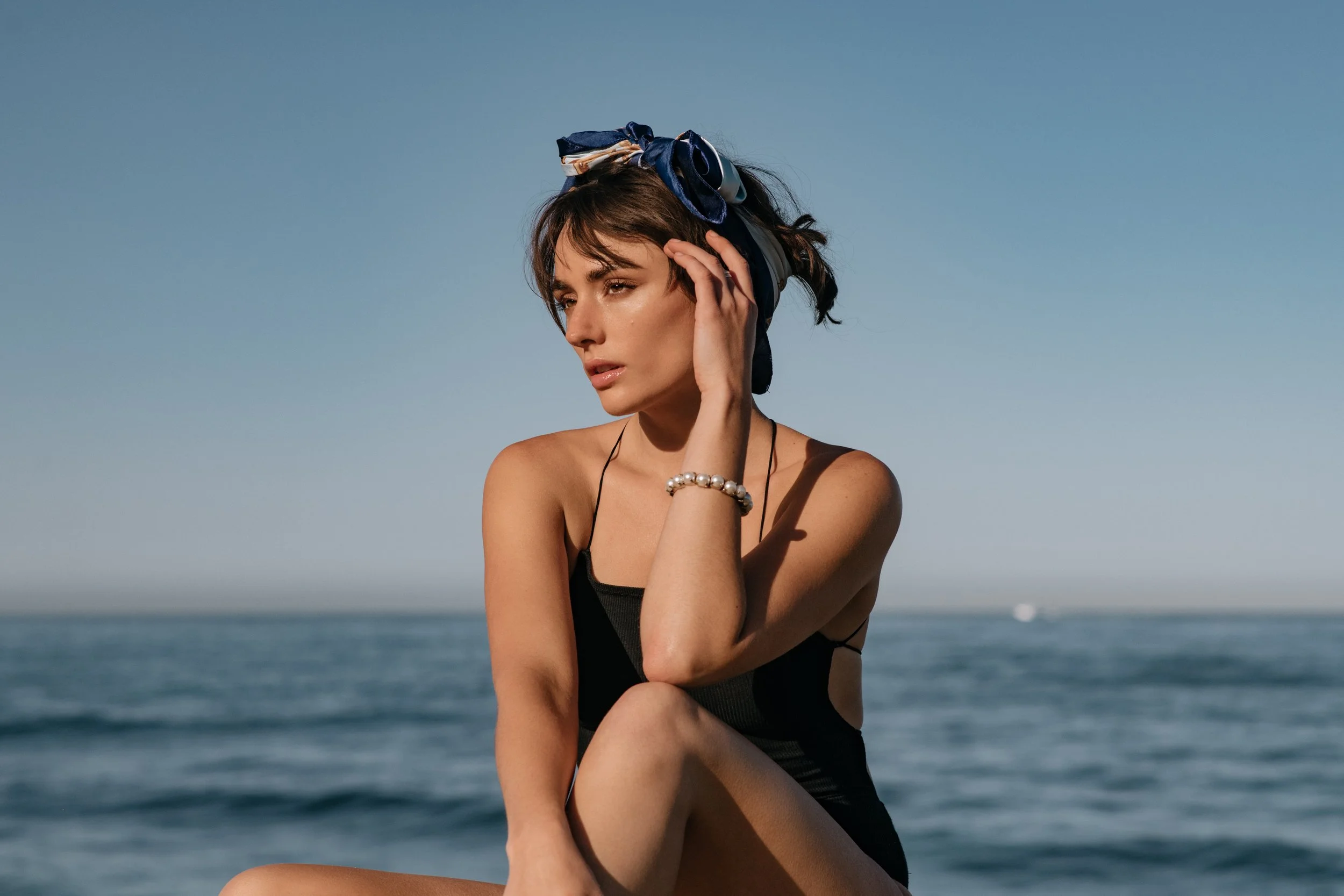 Vintage-inspired swimwear image of model in black one-piece and blue scarf seated by calm ocean 