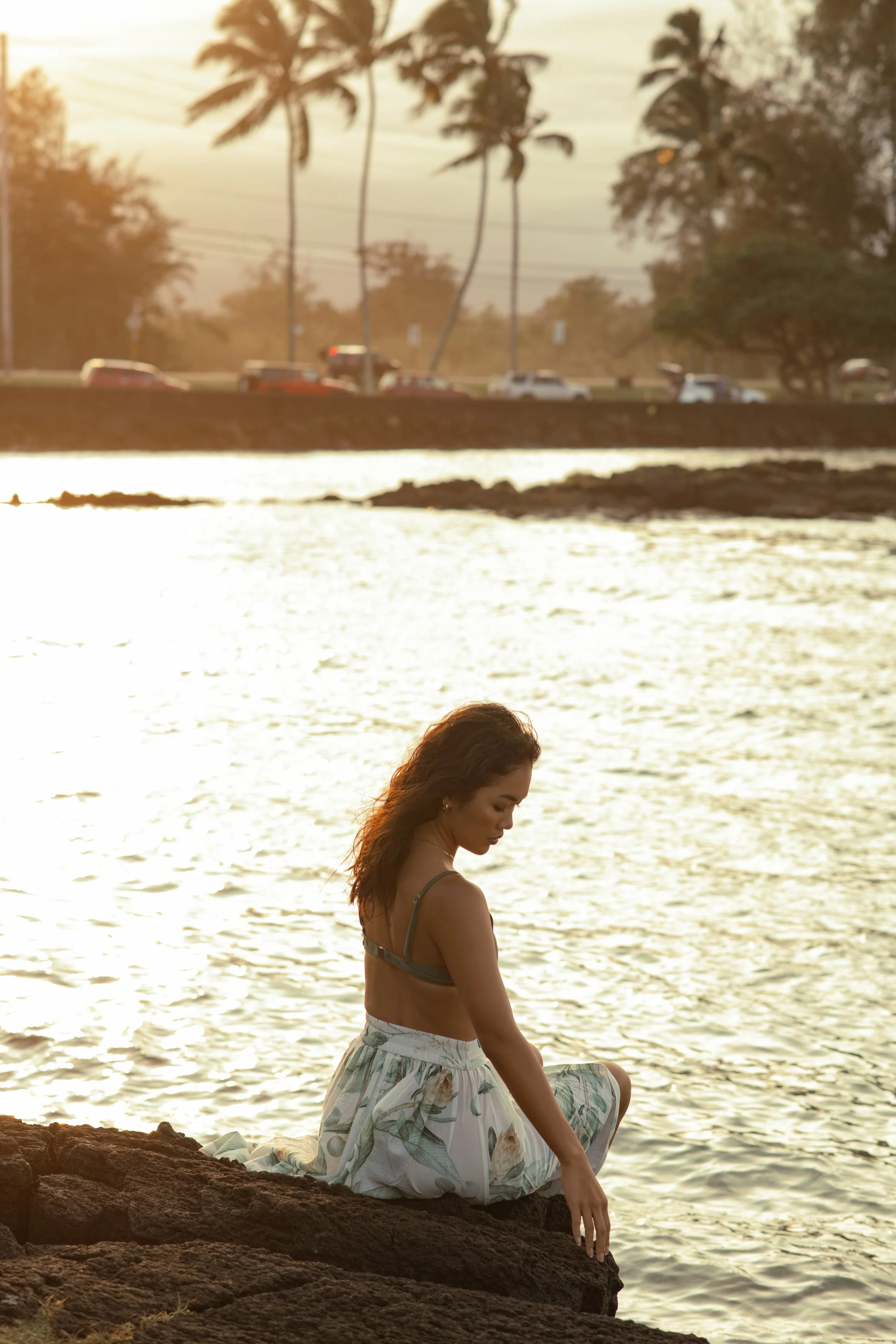  Minimal lifestyle portrait of woman sitting by the ocean at sunset, wearing a flowing white dress in Hawaii 