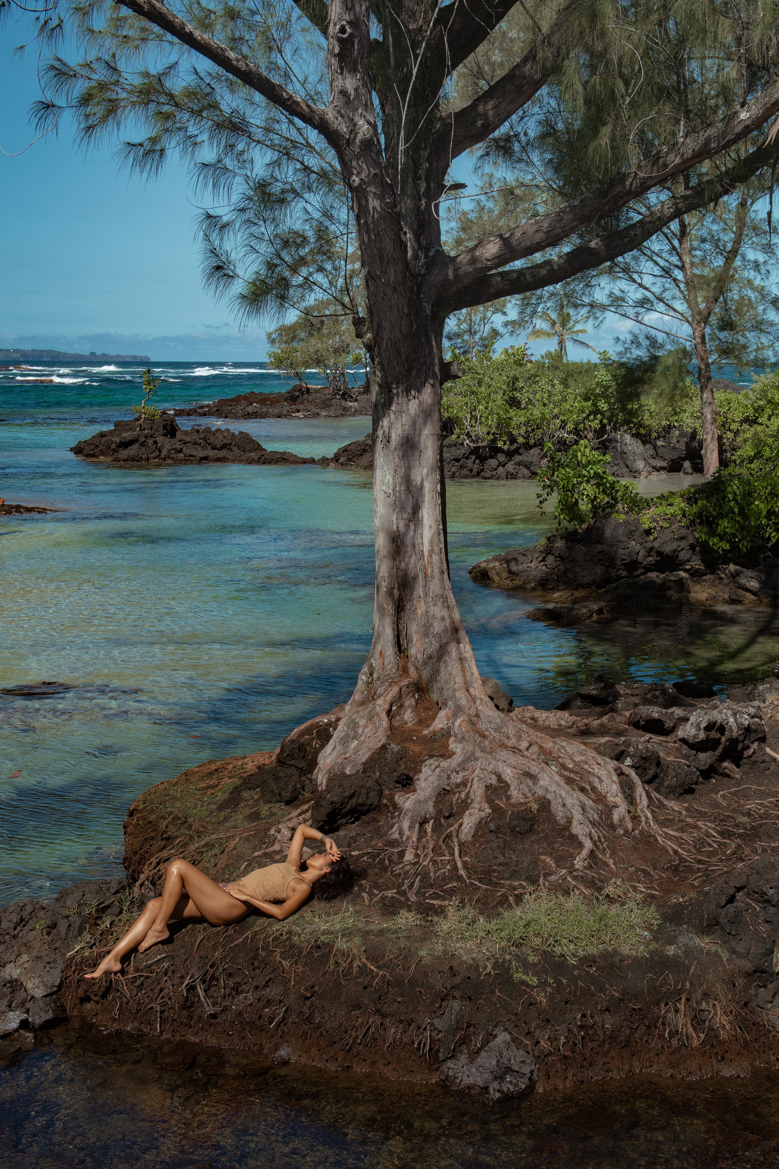  Earthy lifestyle swimwear photo of model resting on volcanic rock under tree with turquoise water in background 