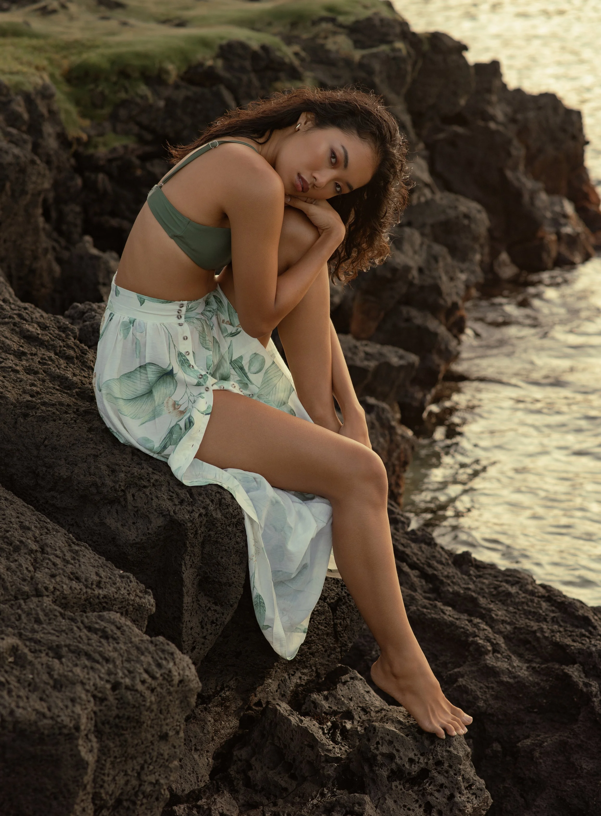  Editorial beachside portrait of model in green bikini and printed skirt, seated on volcanic rocks 