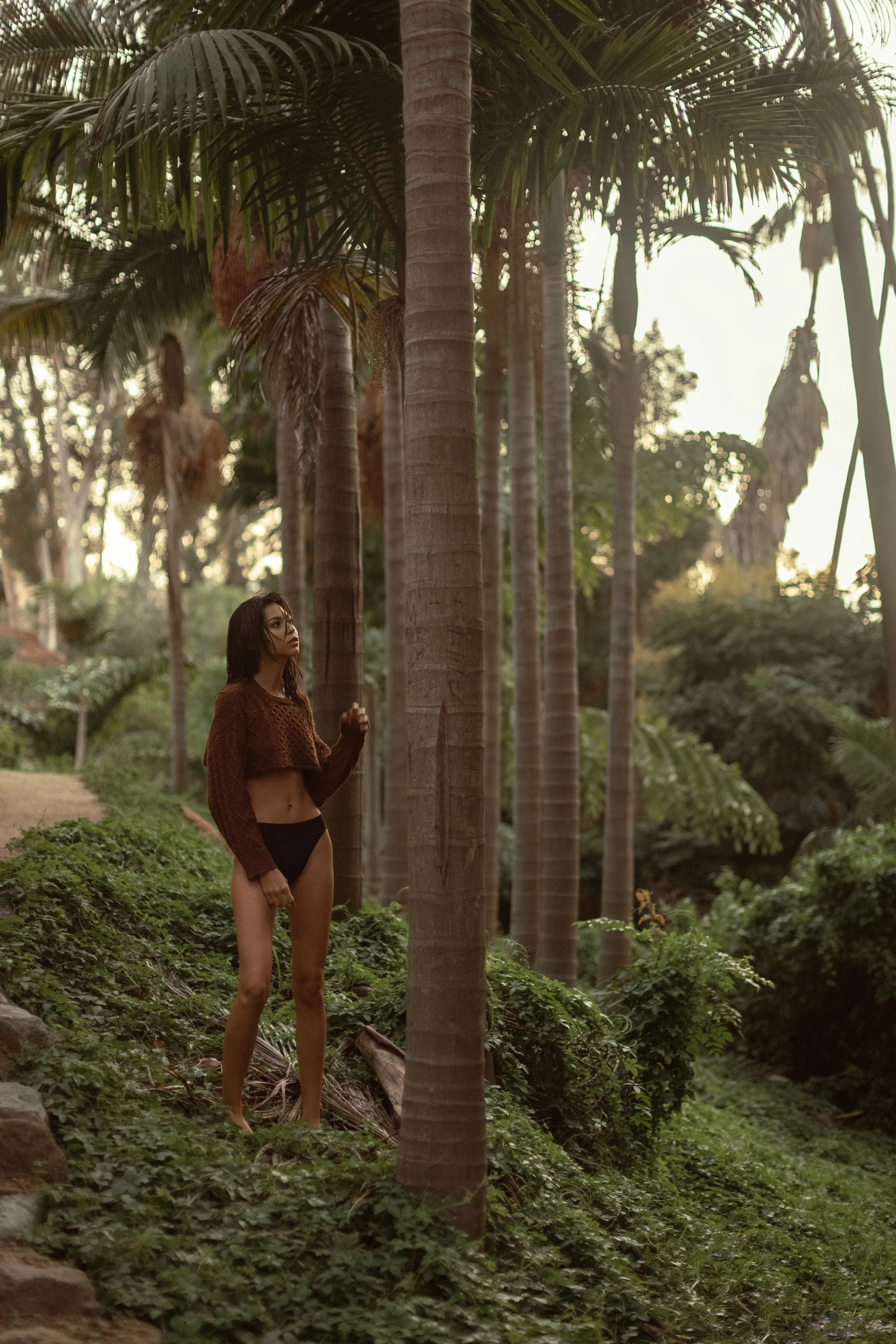  Moody fashion and swimwear image of model standing barefoot in lush, palm-lined jungle setting 