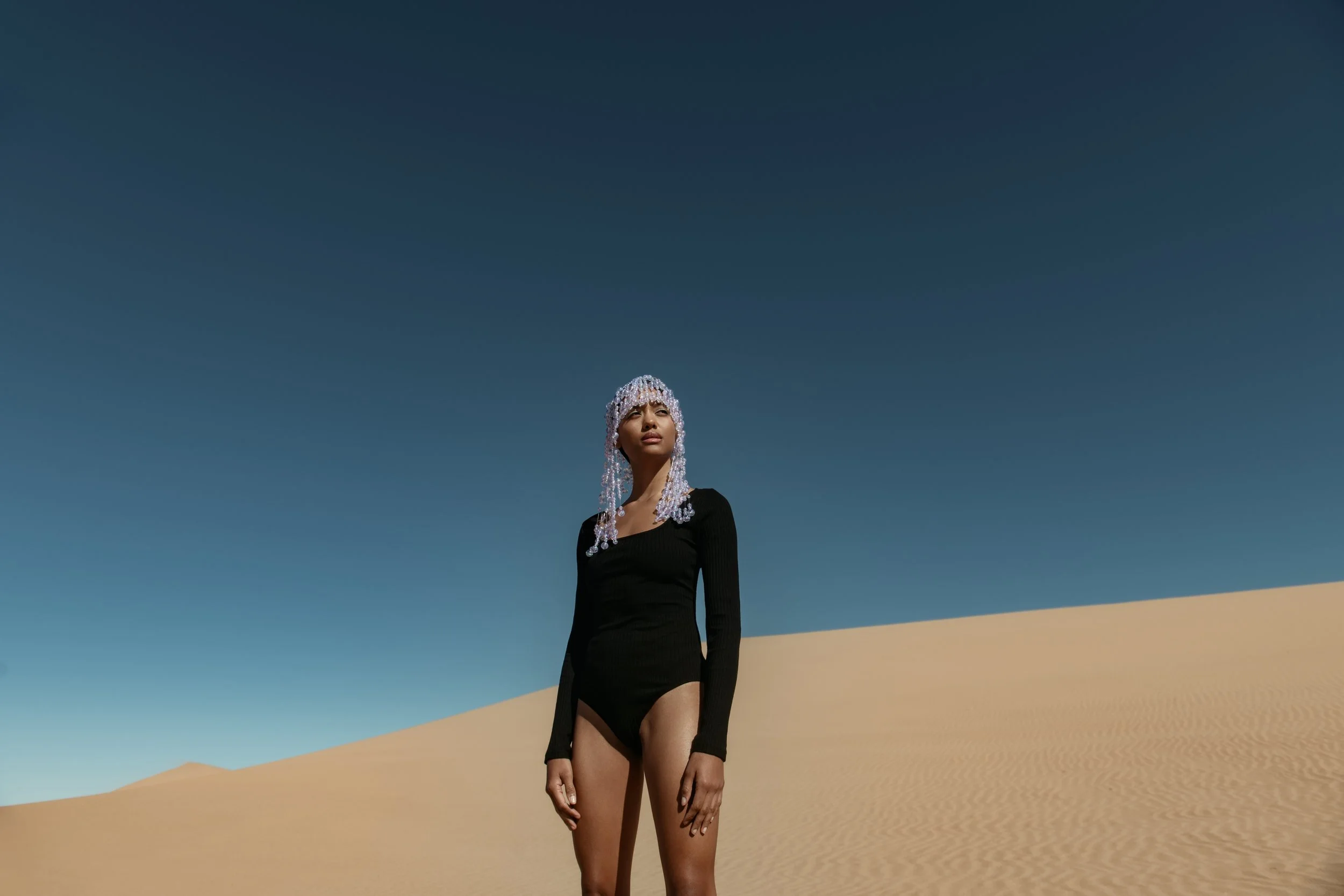  Conceptual fashion portrait of model in black bodysuit and pearl headpiece against sand dunes and blue sky 