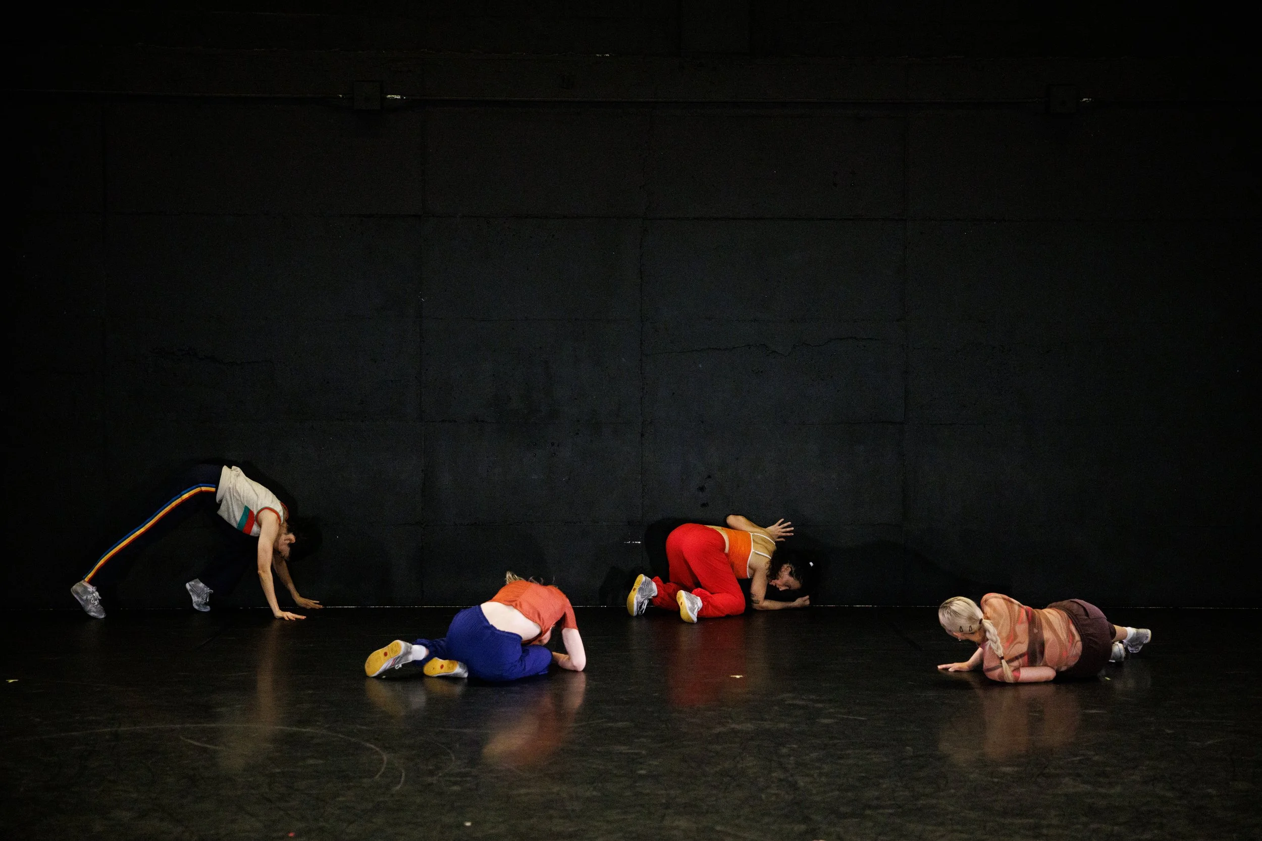 Children in colorful clothing practicing gymnastics or dance on a dark stage with a black wall background.