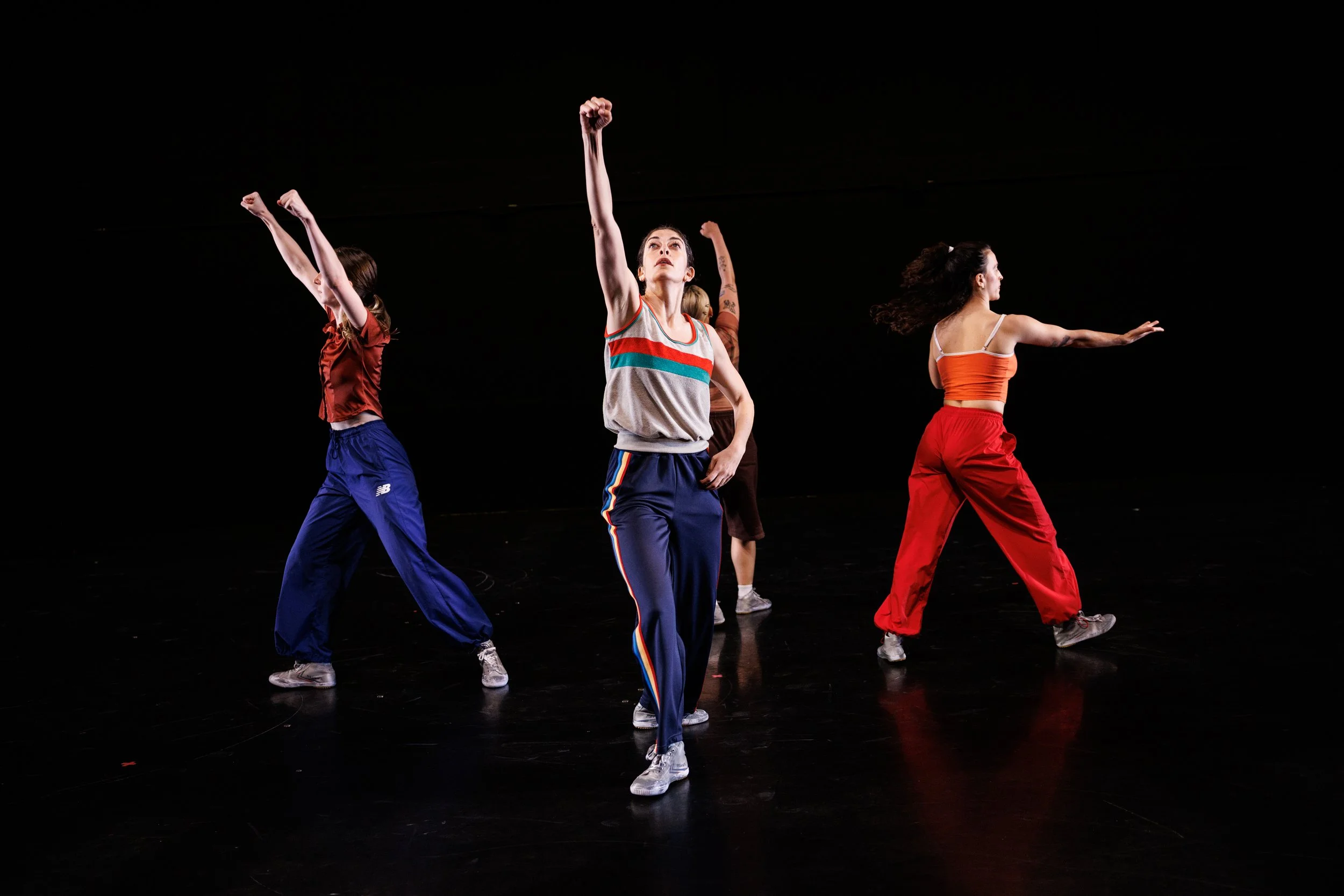 A group of five women dancing on a dark stage, dressed in colorful athletic clothing, with one woman at the center raising her arm.
