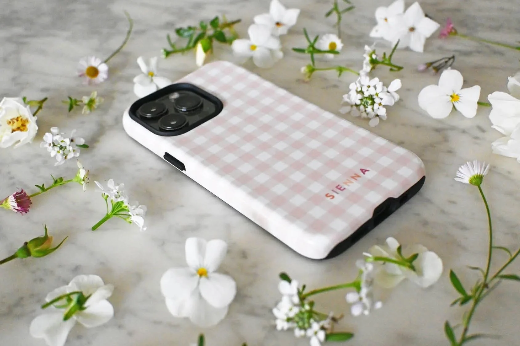 A personalised gingham phone case in blush pink. Personalised with the name Sienna, and styled with fresh white flowers on marble.