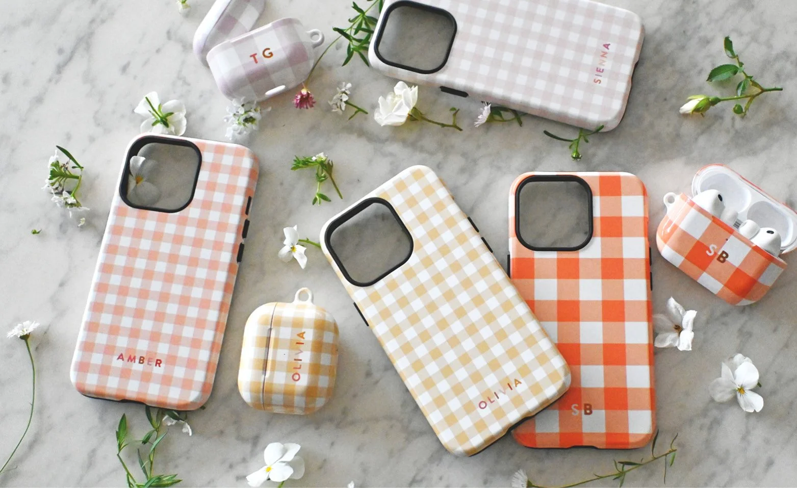 A flat lay of personalised gingham patterned phone and AirPods cases in different warm tones, customised with either names or initials. White flowers are scattered around the custom cases.
