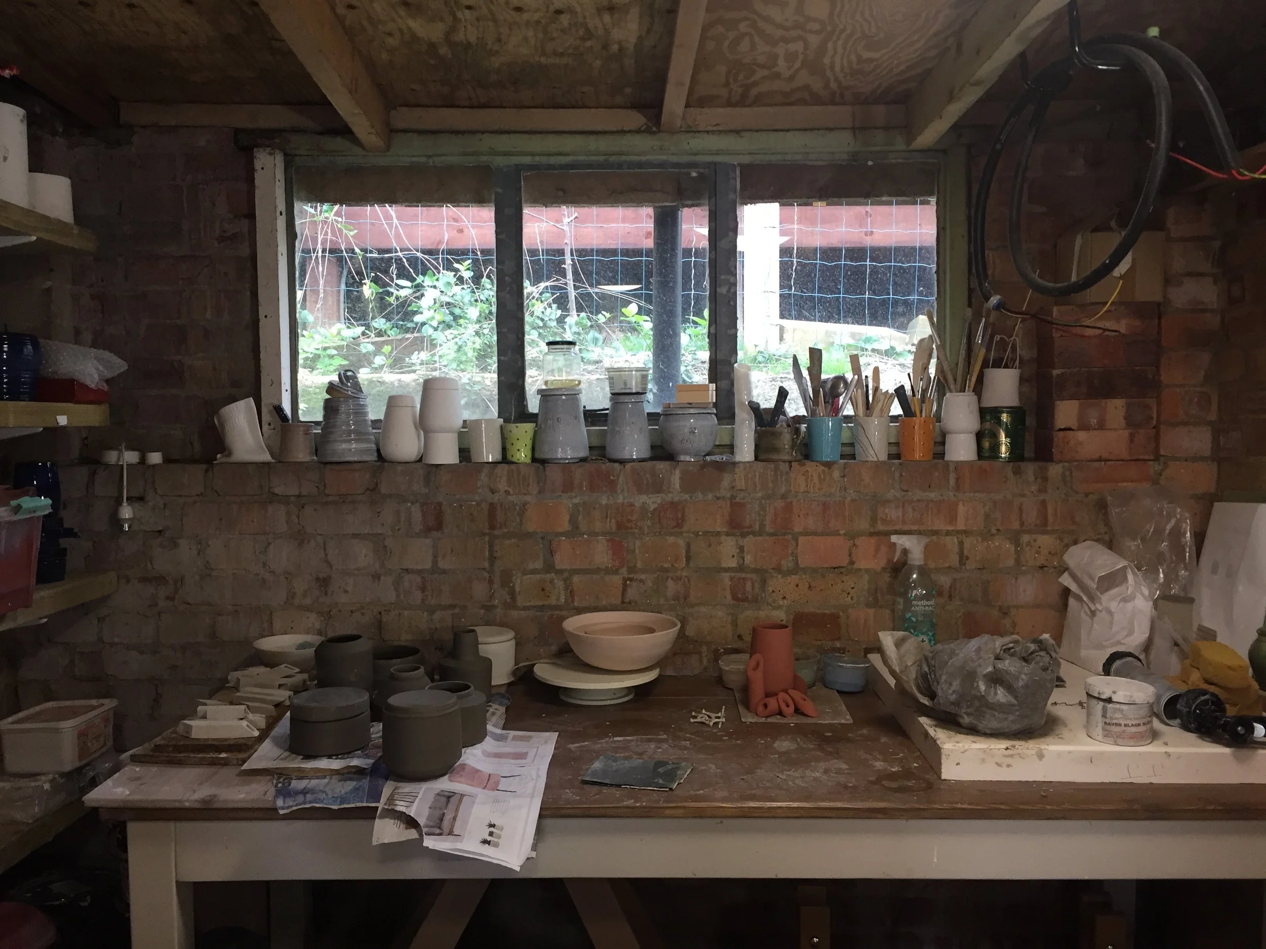 Pottery workspace in north London with various pottery pieces and tools on a wooden table, set against a brick wall with a window