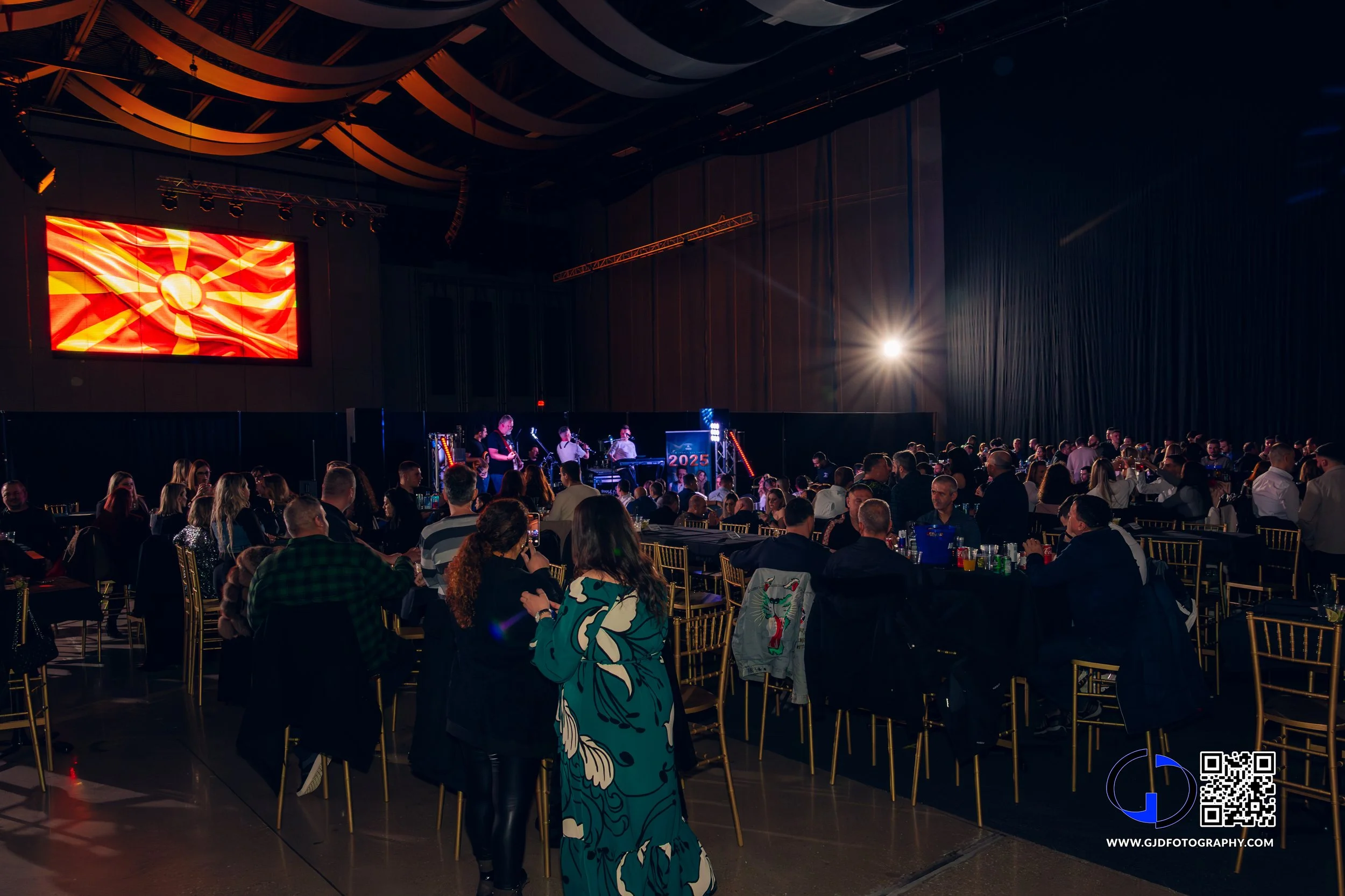 Large indoor event with people sitting at tables watching a stage with a band performing, a large screen displaying the North Macedonian flag, and bright stage lighting.