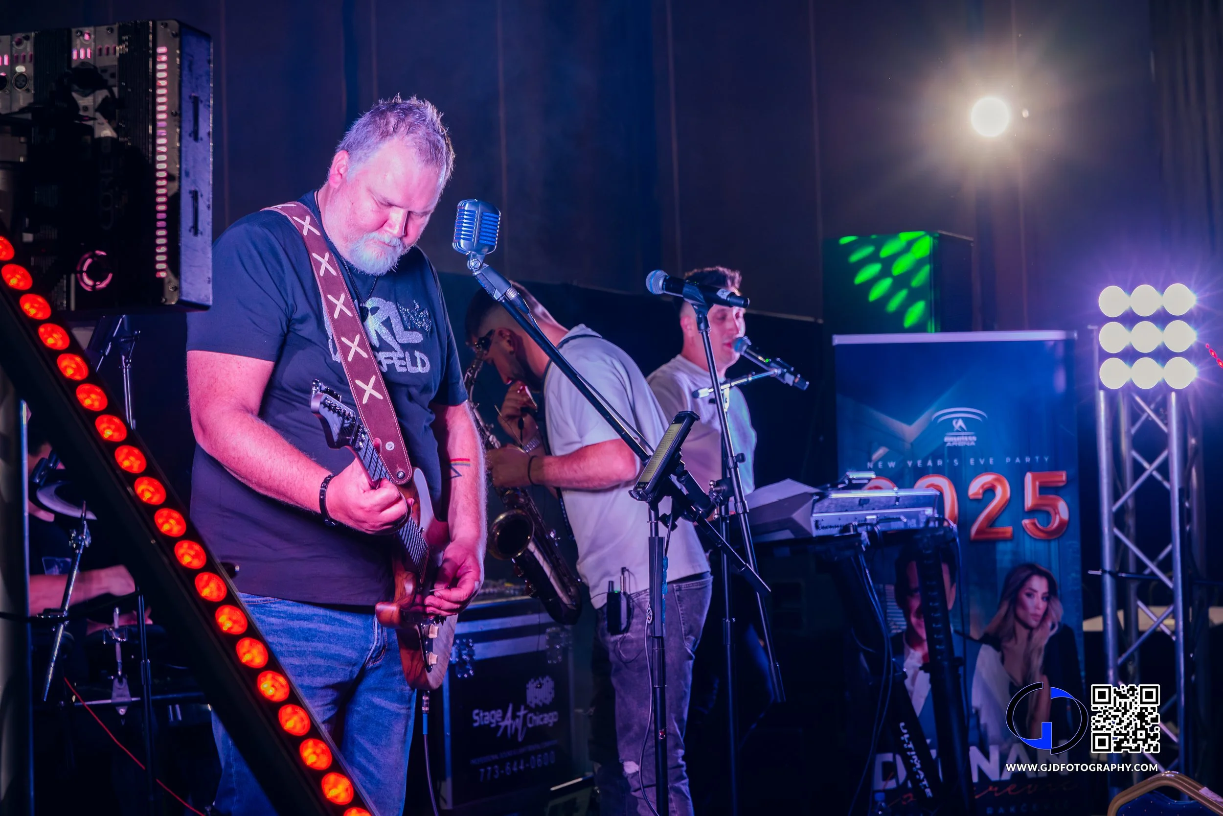 A band performing on stage with members playing guitar, keyboard, and singing into microphones at a New Year's Eve party, with colored stage lights and a large screen displaying the number 2025.