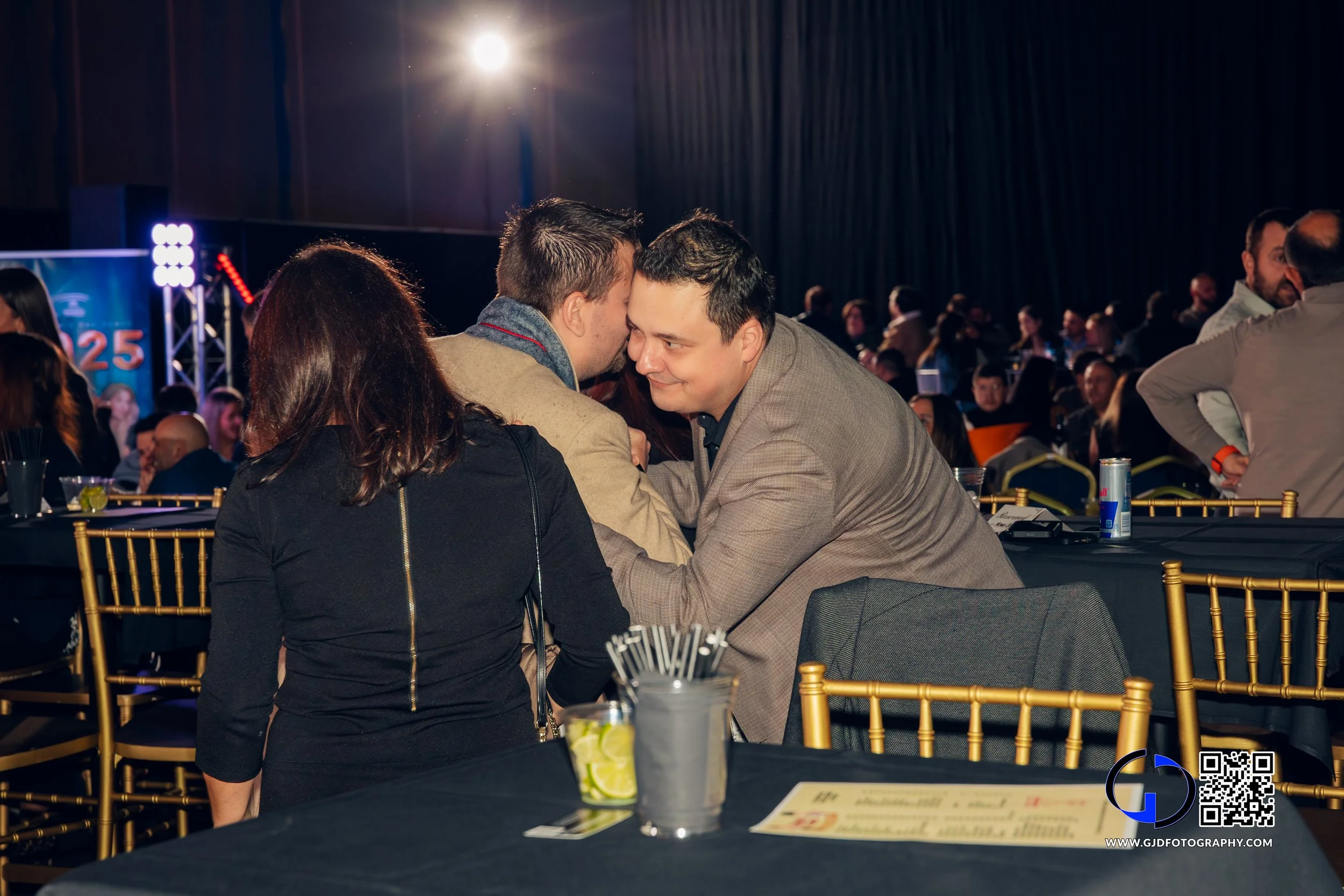 Two men in suits shaking hands and leaning in close at a social event, with a woman in a black jacket observing, in a crowded indoor venue with dim lighting and a stage in the background.