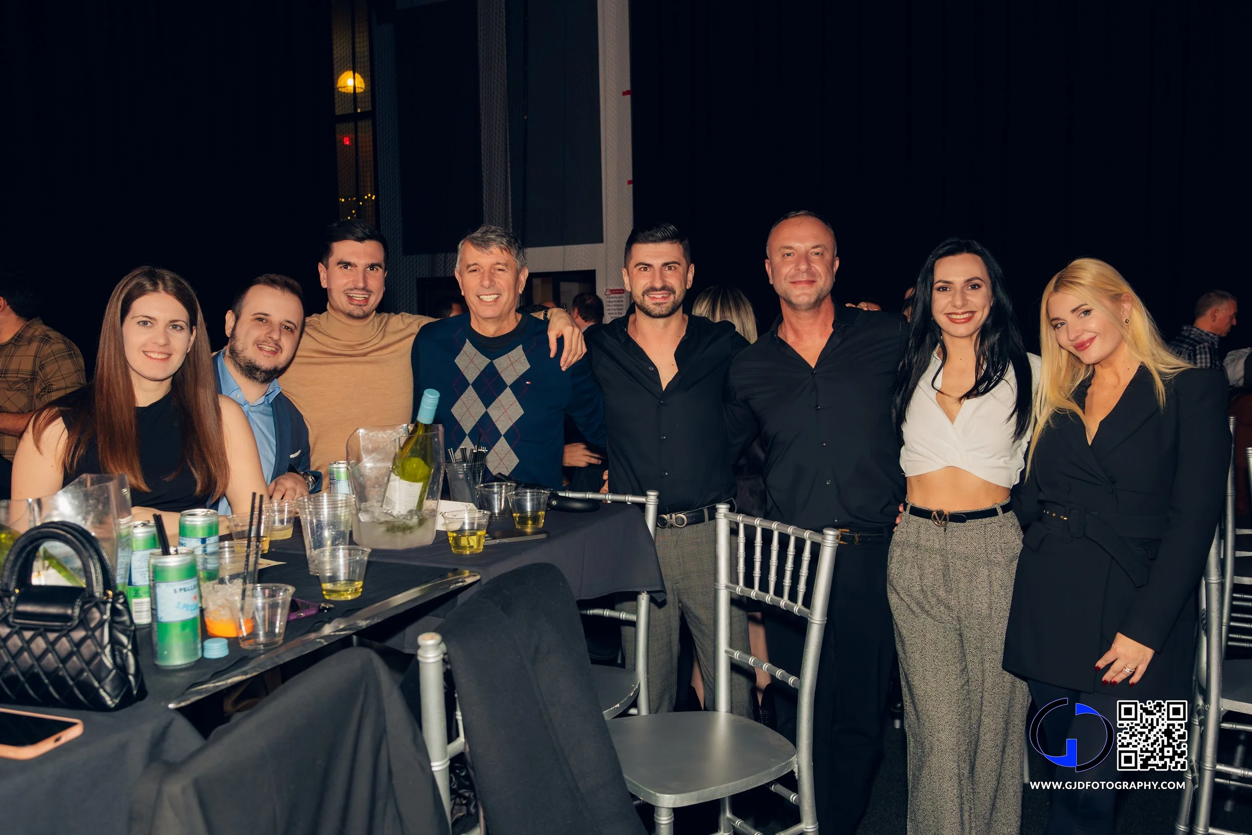 Group of eight people smiling and posing together in a semi-formal setting at a social gathering or party with tables, drinks, and a dark background.