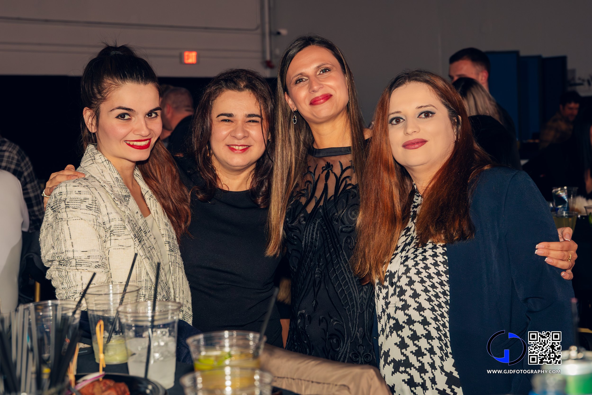 Four women posing for a photo at a social gathering, smiling and standing close together. Drinks and table items are visible in the foreground.