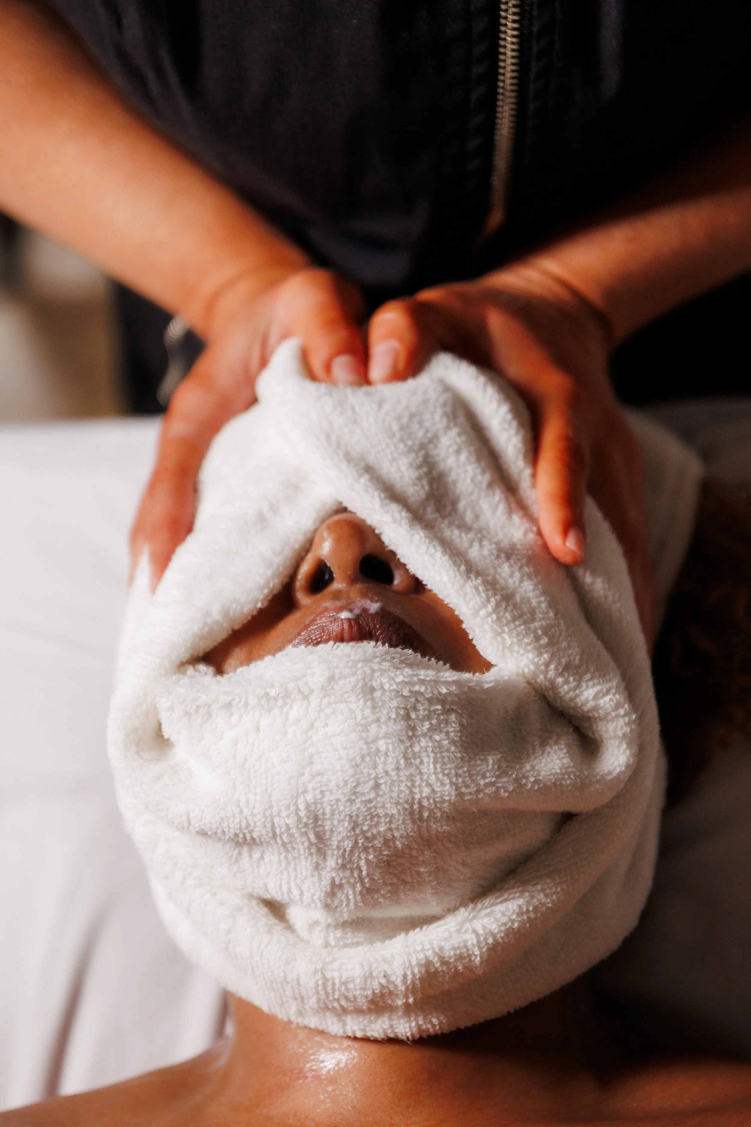 Person receiving a facial treatment with a white towel covering their face, lying down with a technician's hands on their face.
