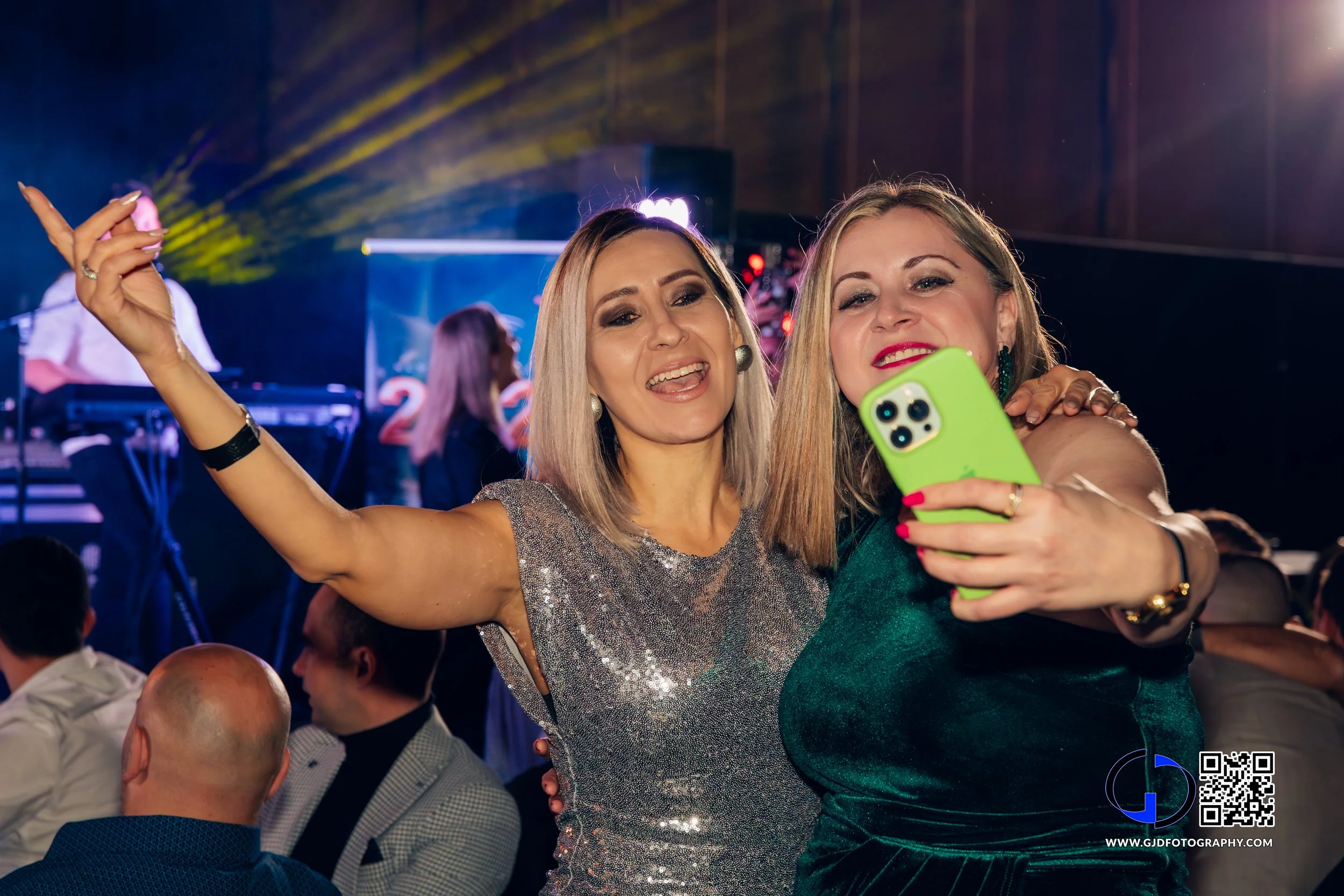 Two women taking a selfie at a party or celebration with a live band and flashing lights in the background.