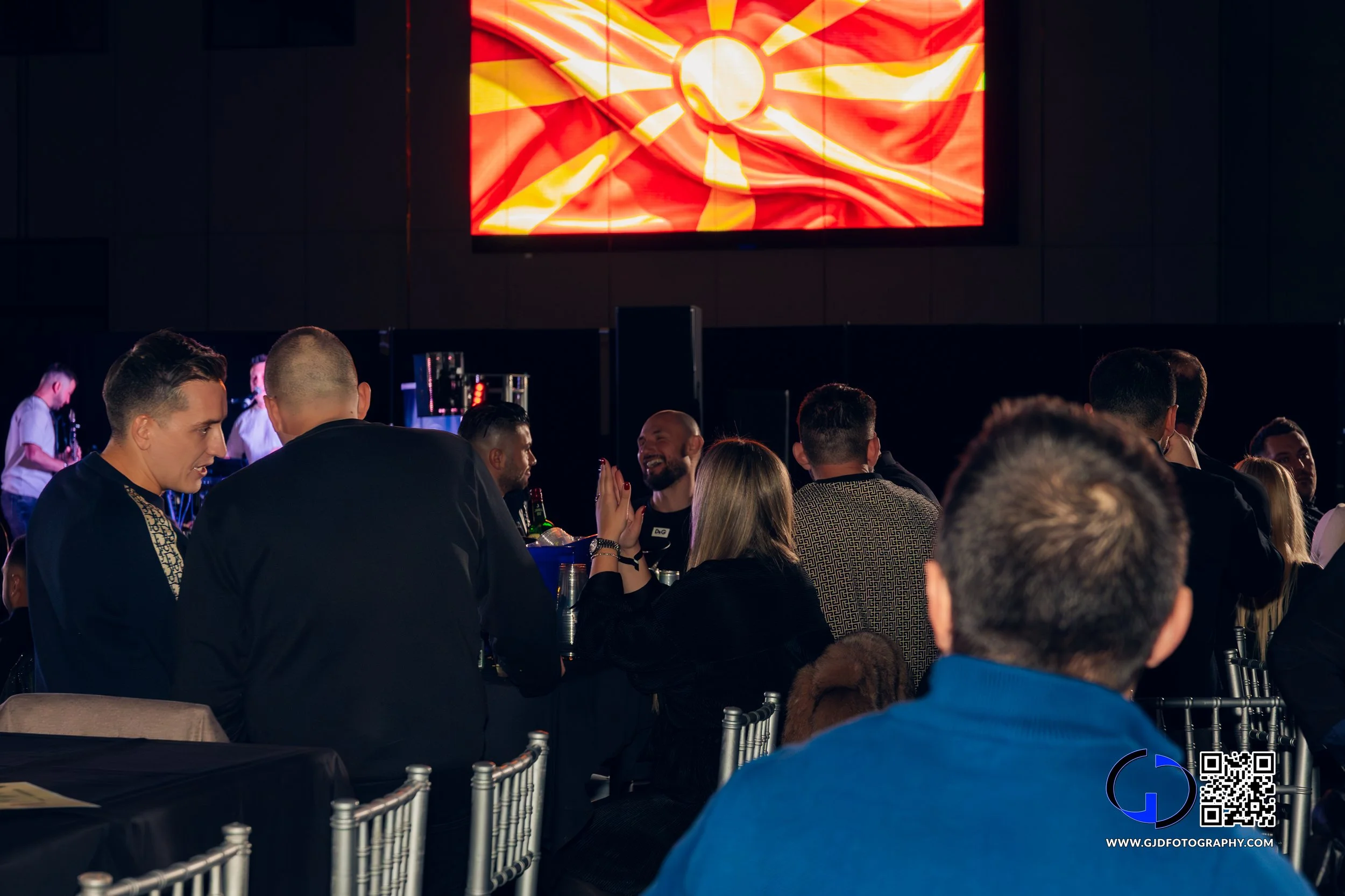 People sitting at tables in a dark room with a large screen displaying a stylized sun and flag design.
