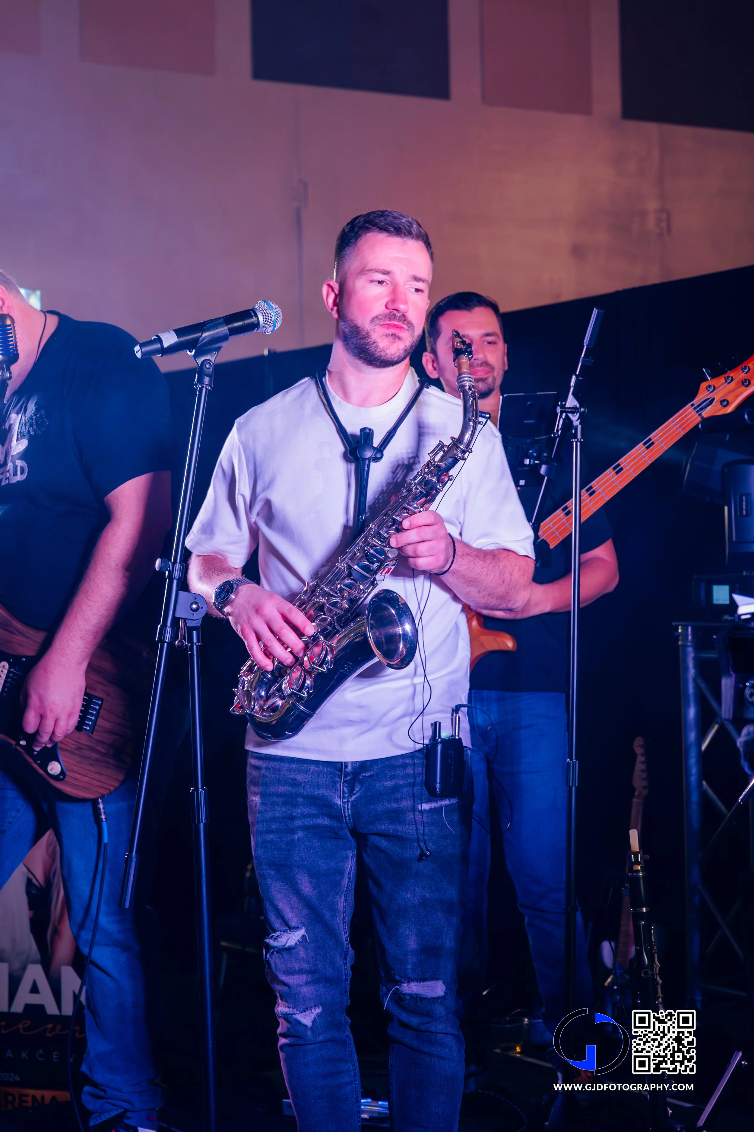 Musician playing saxophone during a live performance, standing in front of microphone with other band members playing guitar and bass in the background.