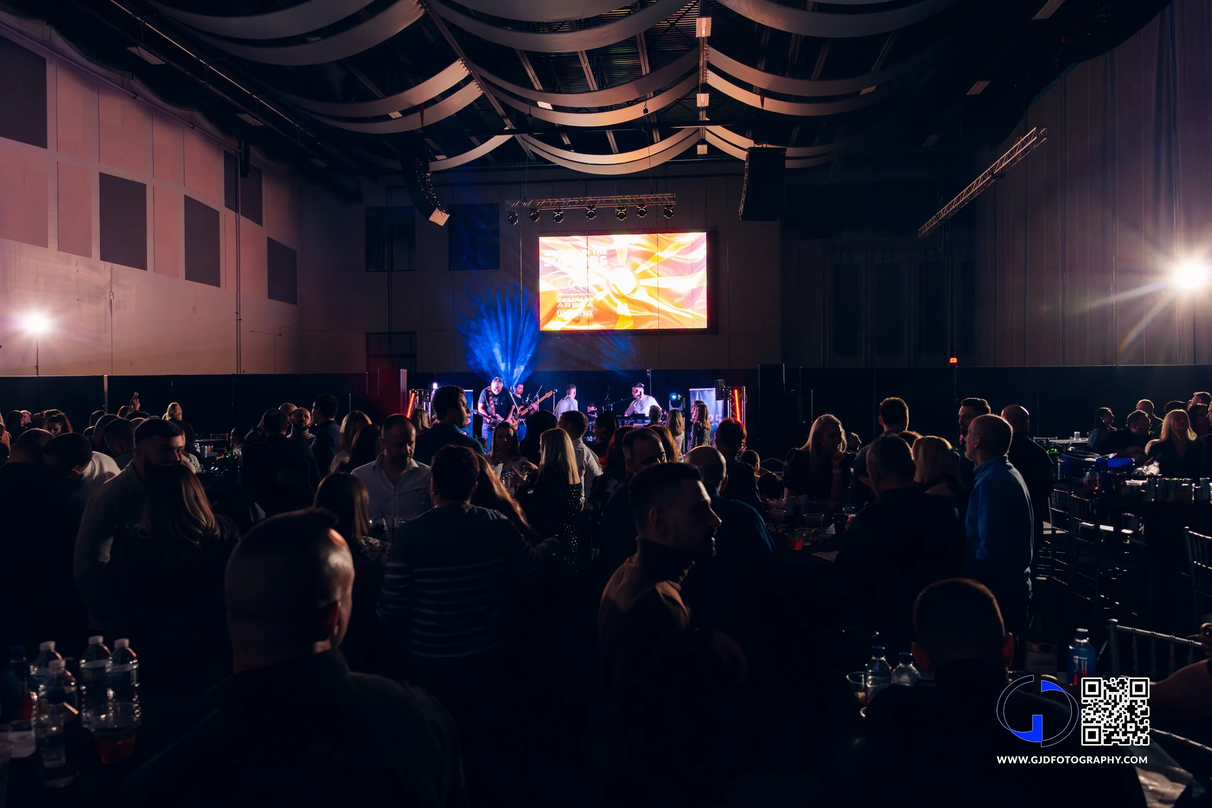 Concert or event with a band performing on stage, audience seated at tables in a large indoor venue with a digital screen and lighting.