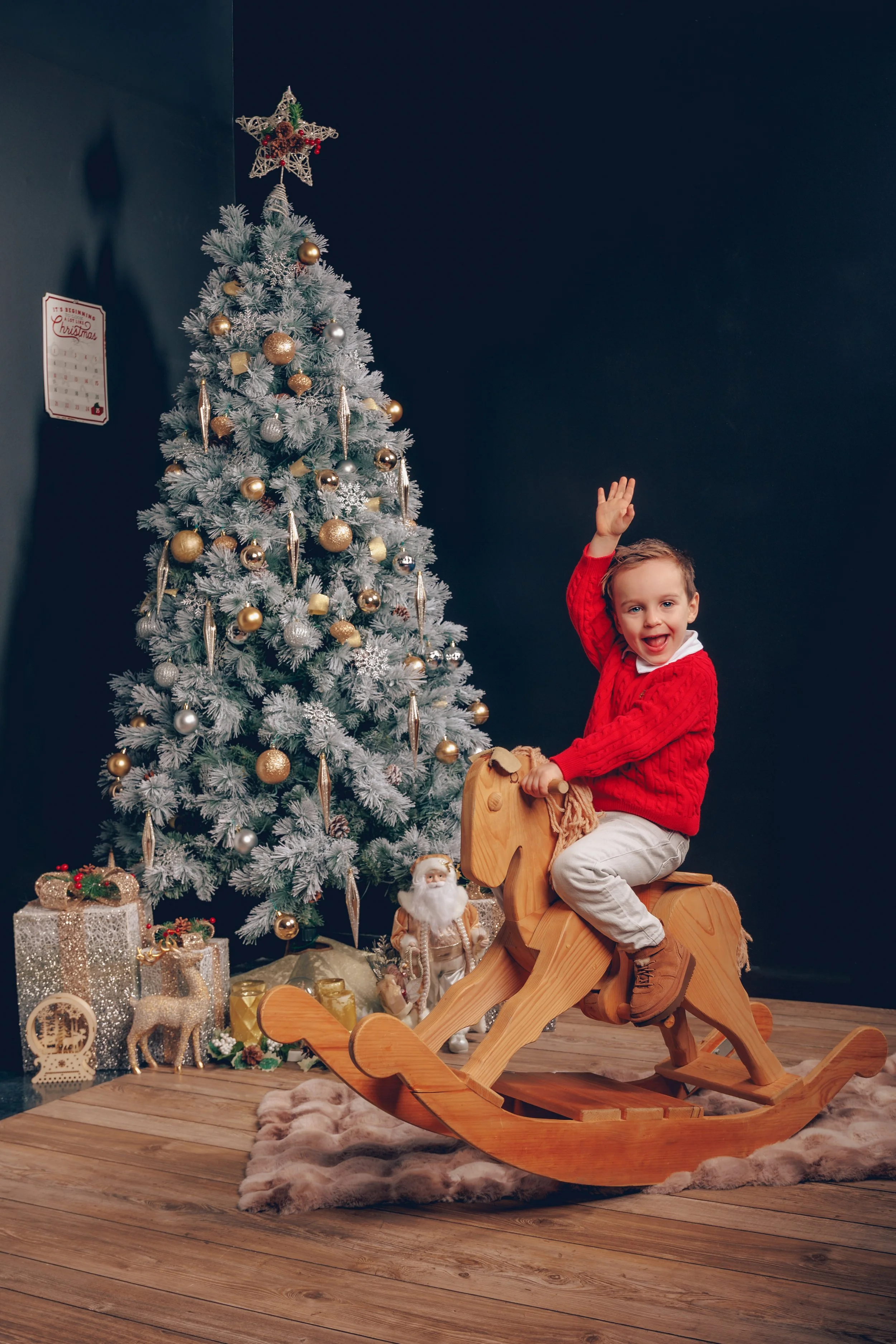A young boy in a red sweater riding a wooden rocking horse in front of a decorated Christmas tree with presents.