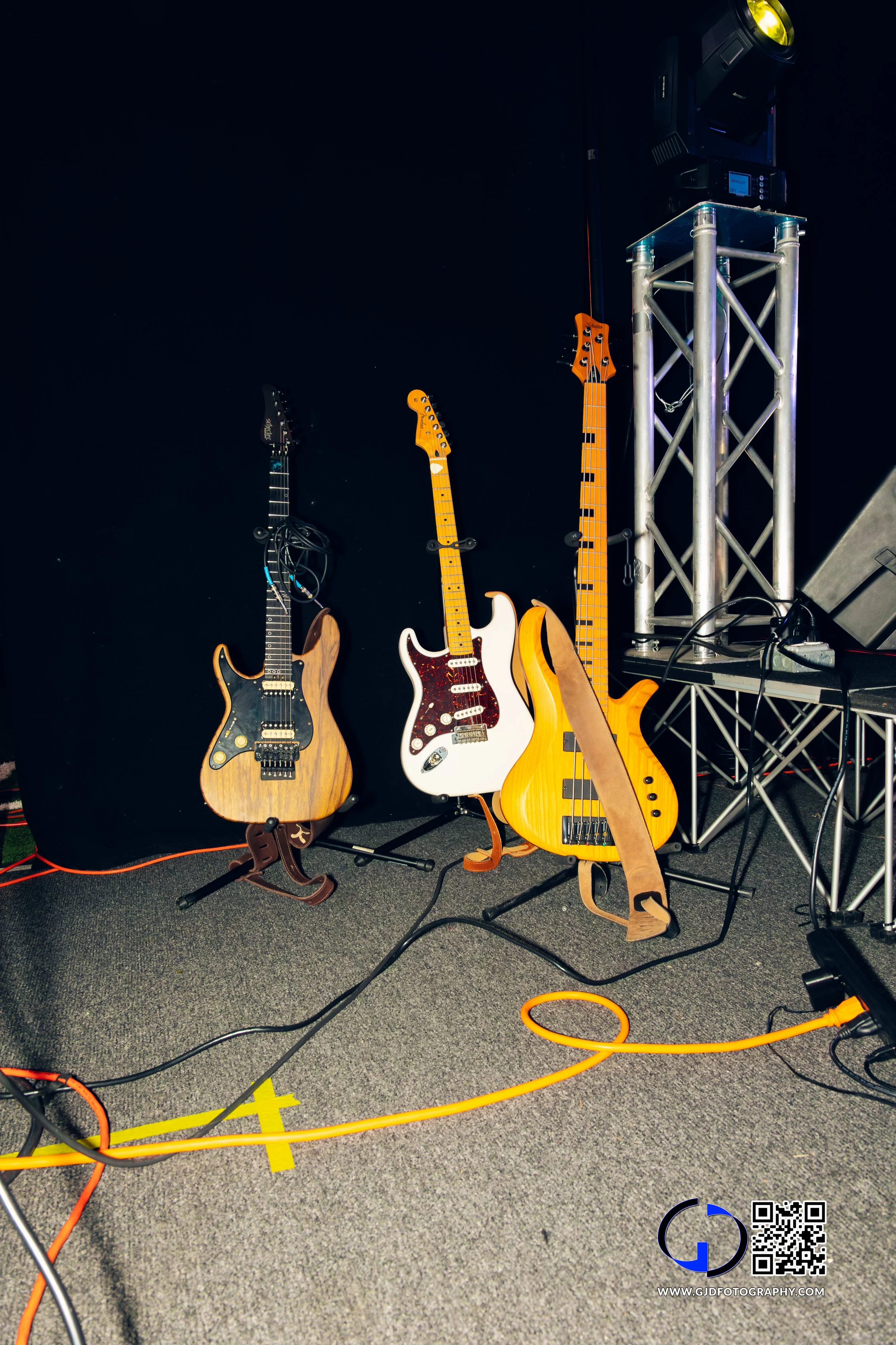Three electric guitars on stands, set against a black background, with sound equipment and cables on a grey carpeted floor.