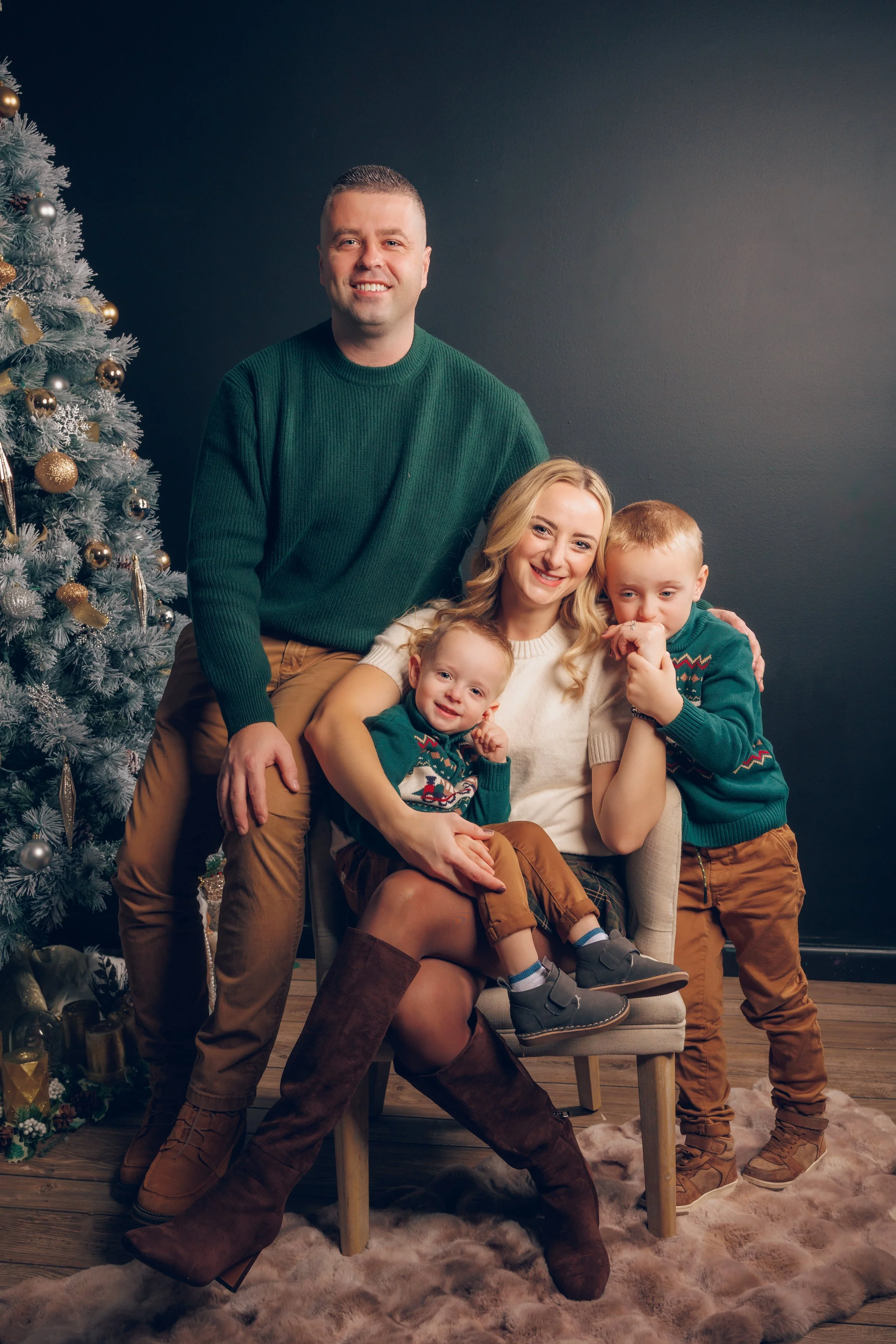 A family of four posing near a decorated Christmas tree. The father is standing, wearing a green sweater. The mother is sitting on a chair with two children, both wearing matching green holiday sweaters. The scene is cozy with a dark wall background 