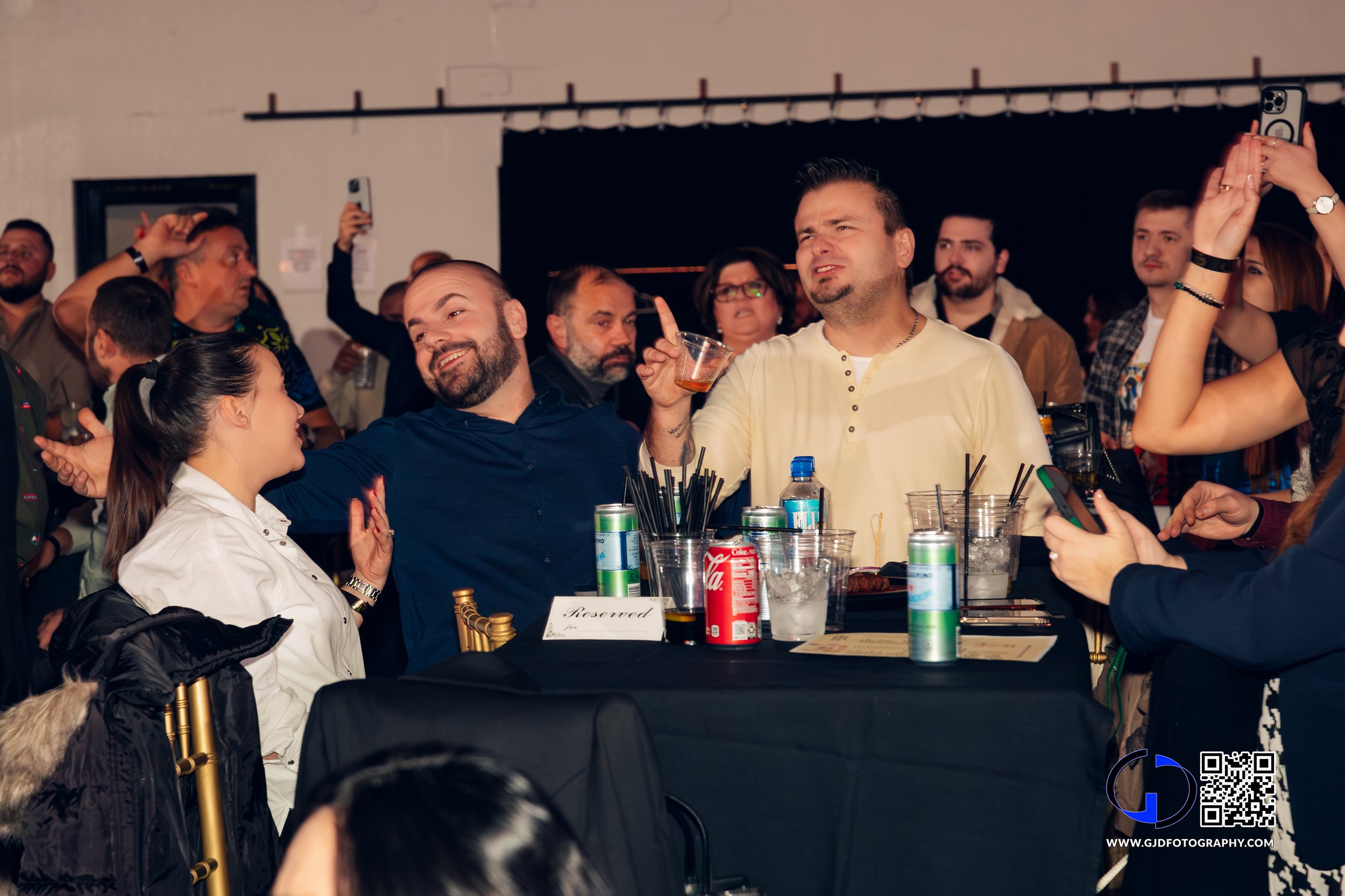 People at a social event, some speaking and others taking photos, with drinks and snacks on the table.
