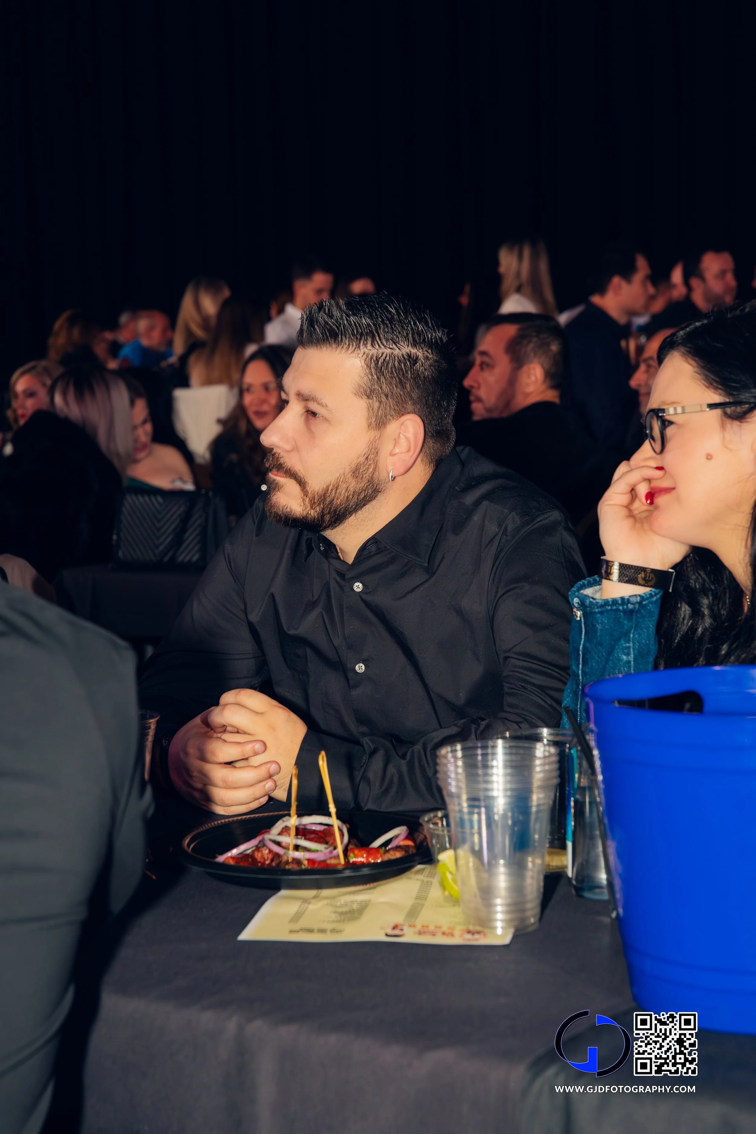A man with dark hair and beard wearing a black shirt, sitting at a table during an event, with a plate of food in front of him and a woman with dark hair and glasses sitting nearby. In the background, multiple people are seated at tables, some engage