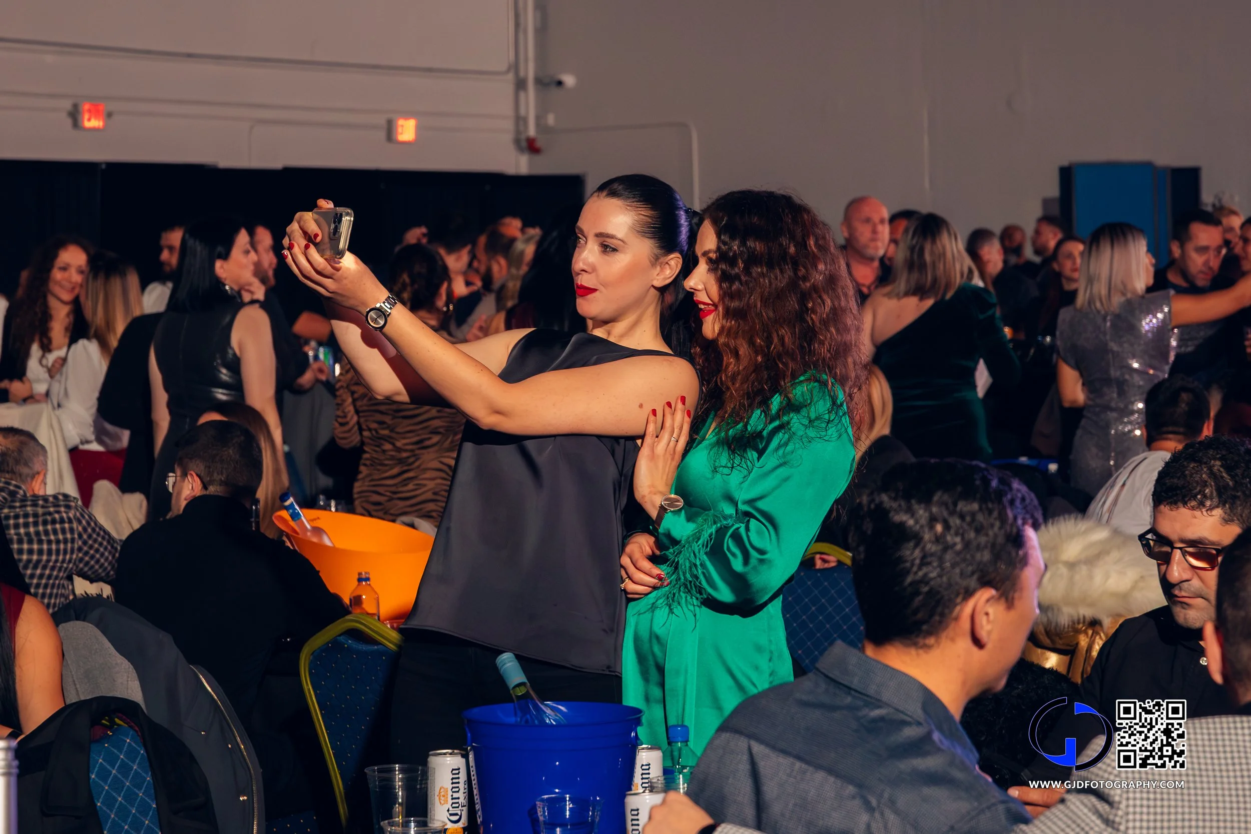 Two women taking a selfie at a crowded indoor social event, with tables, drinks, and numerous people in the background.