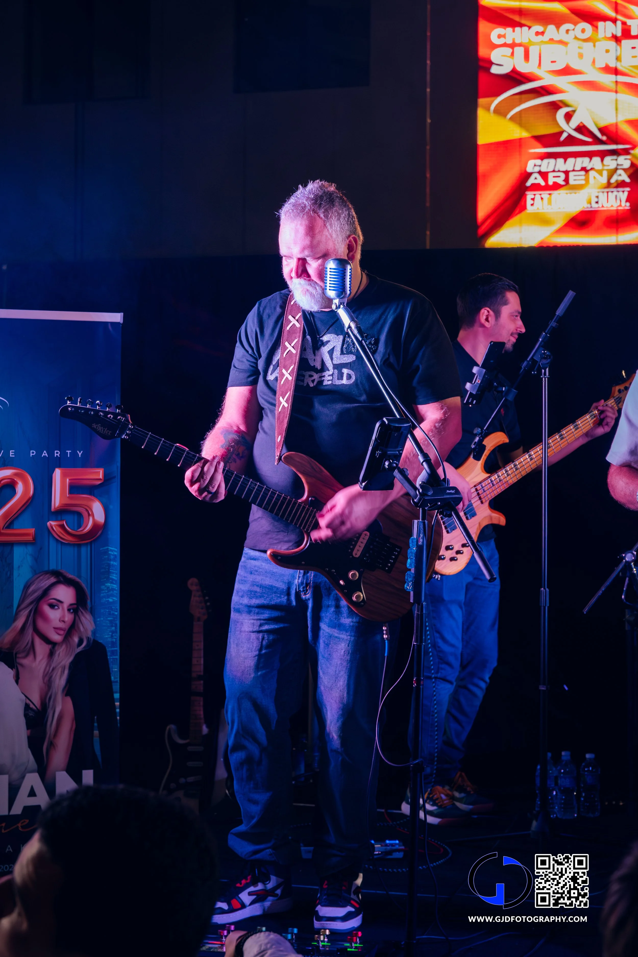 Male guitarist with gray beard playing electric guitar on stage, with a microphone in front of him and another musician in the background playing bass guitar, illuminated by stage lights.
