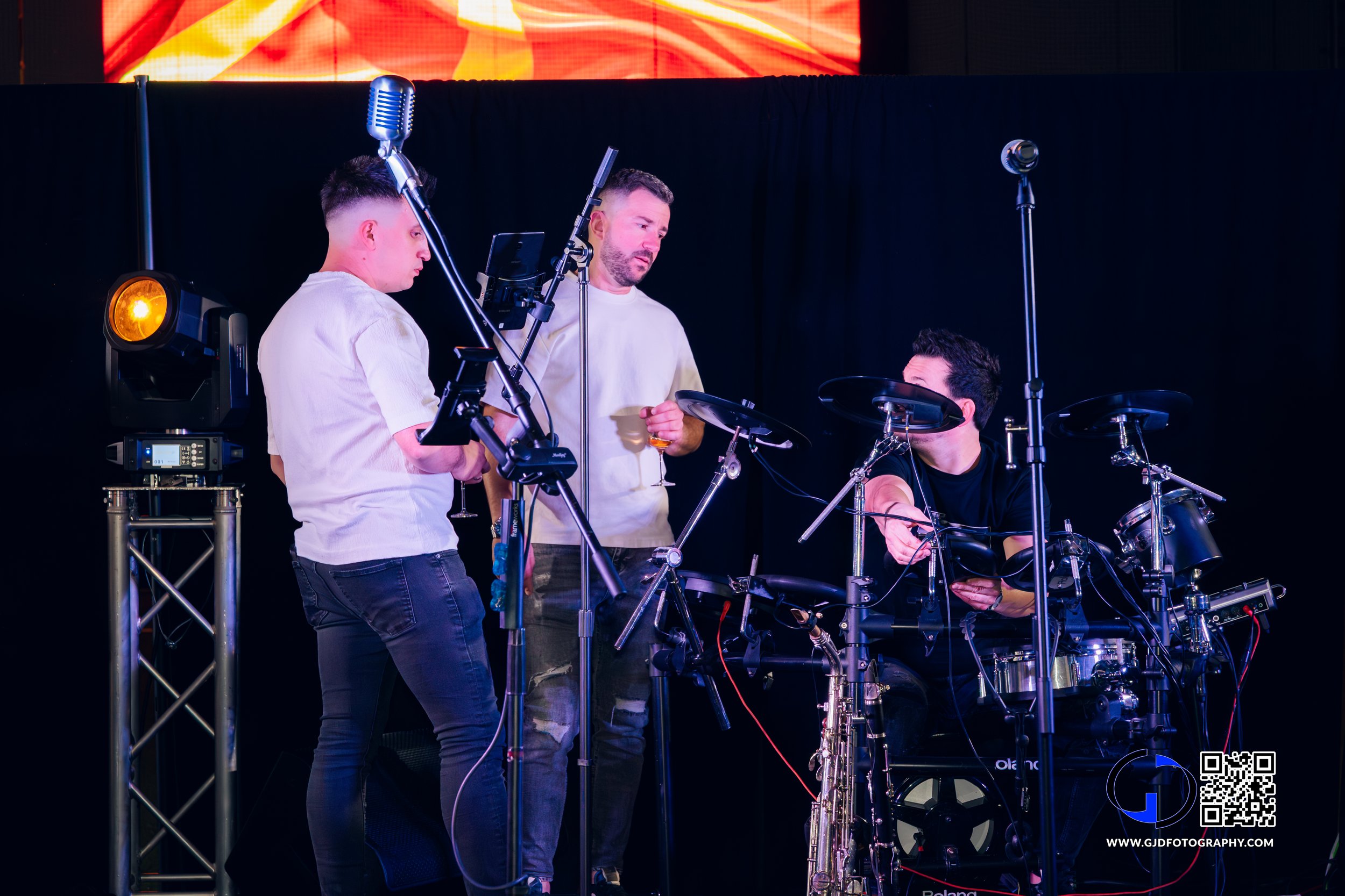 Three men are working on a digital drum set on a stage with a dark backdrop and a colorful digital display above. One man is standing and holding a glass, the other two are seated, one adjusting equipment and the other observing.