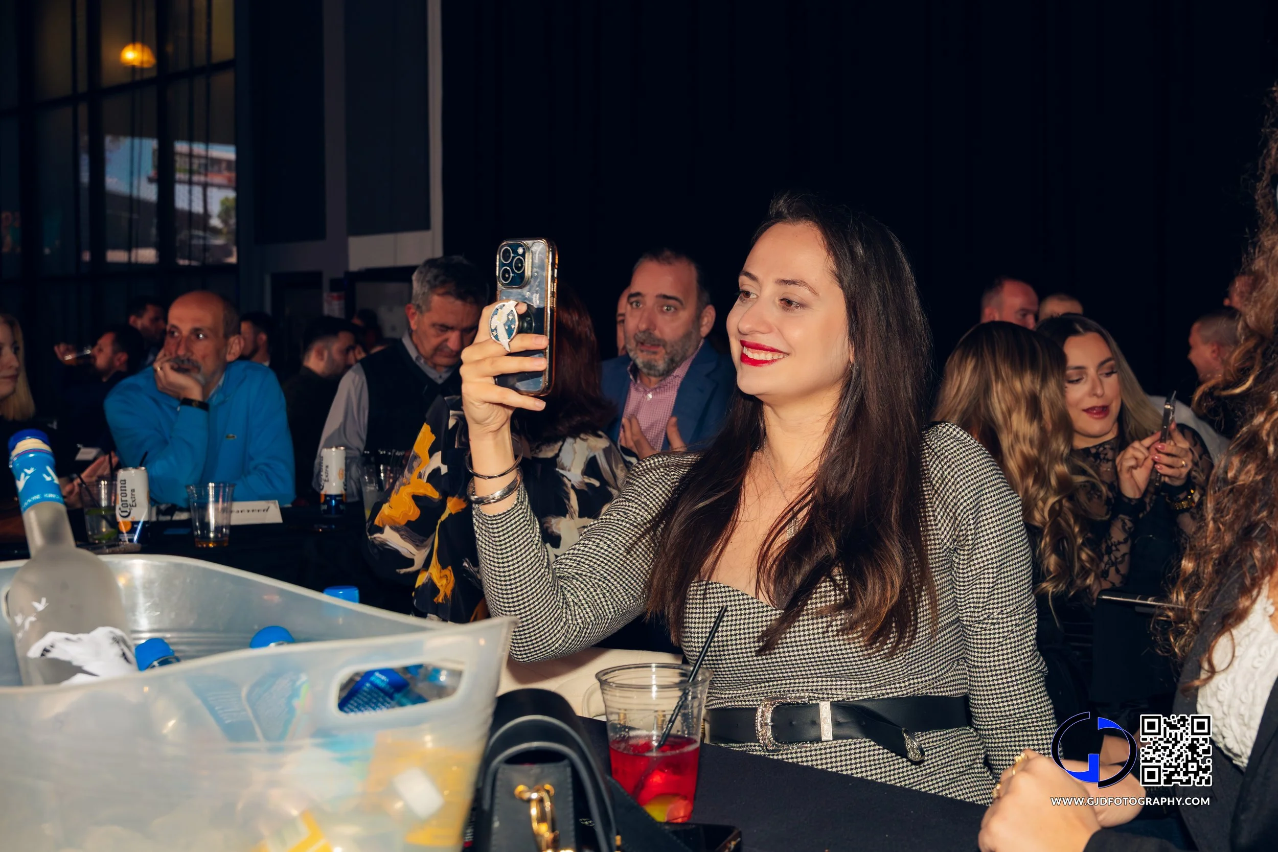 Young woman taking a selfie at a crowded indoor event with beverages on the table.