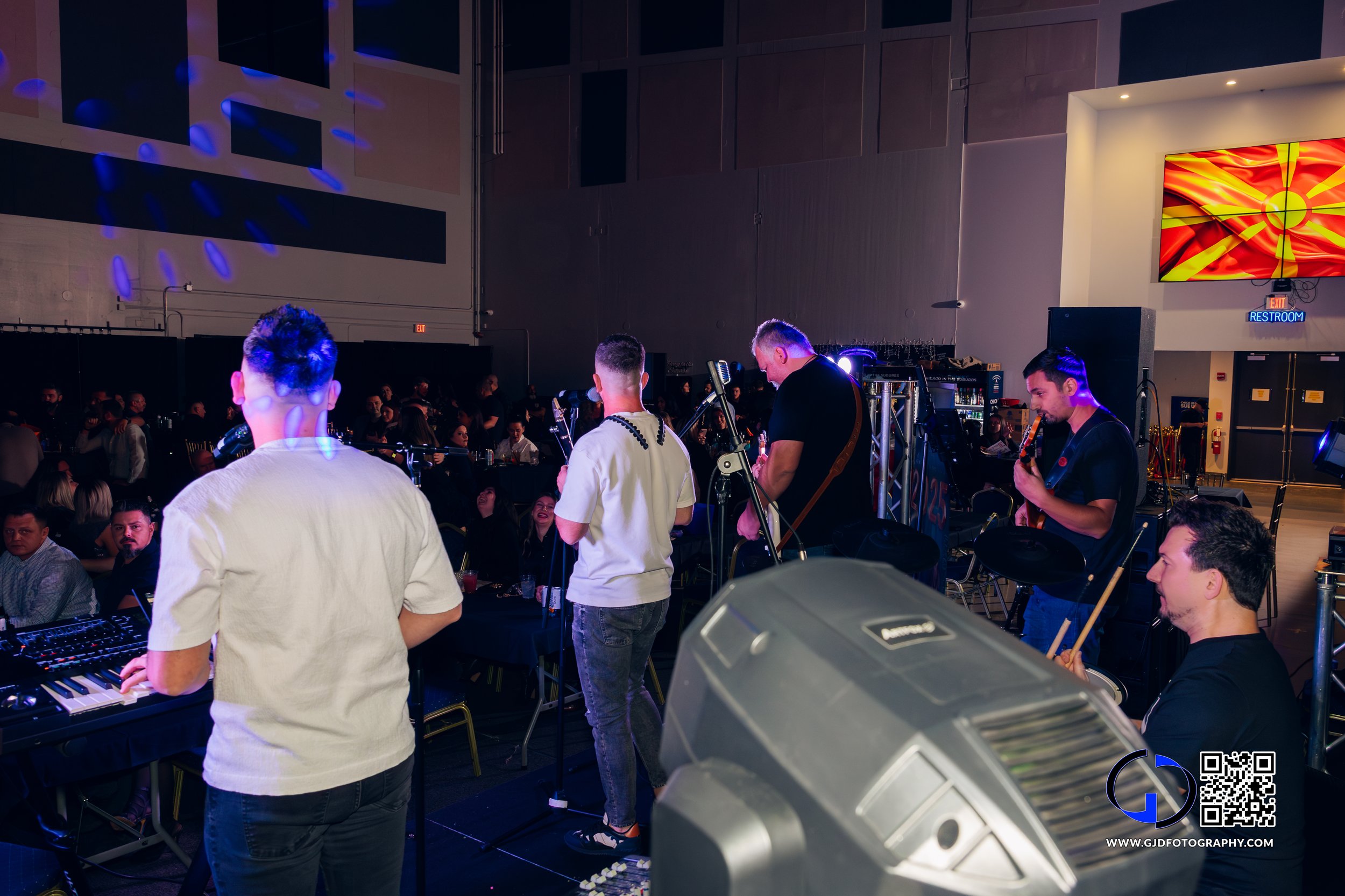 Musicians performing on stage at an indoor event with audience seated at tables, colorful lighting, and a large screen in the background.