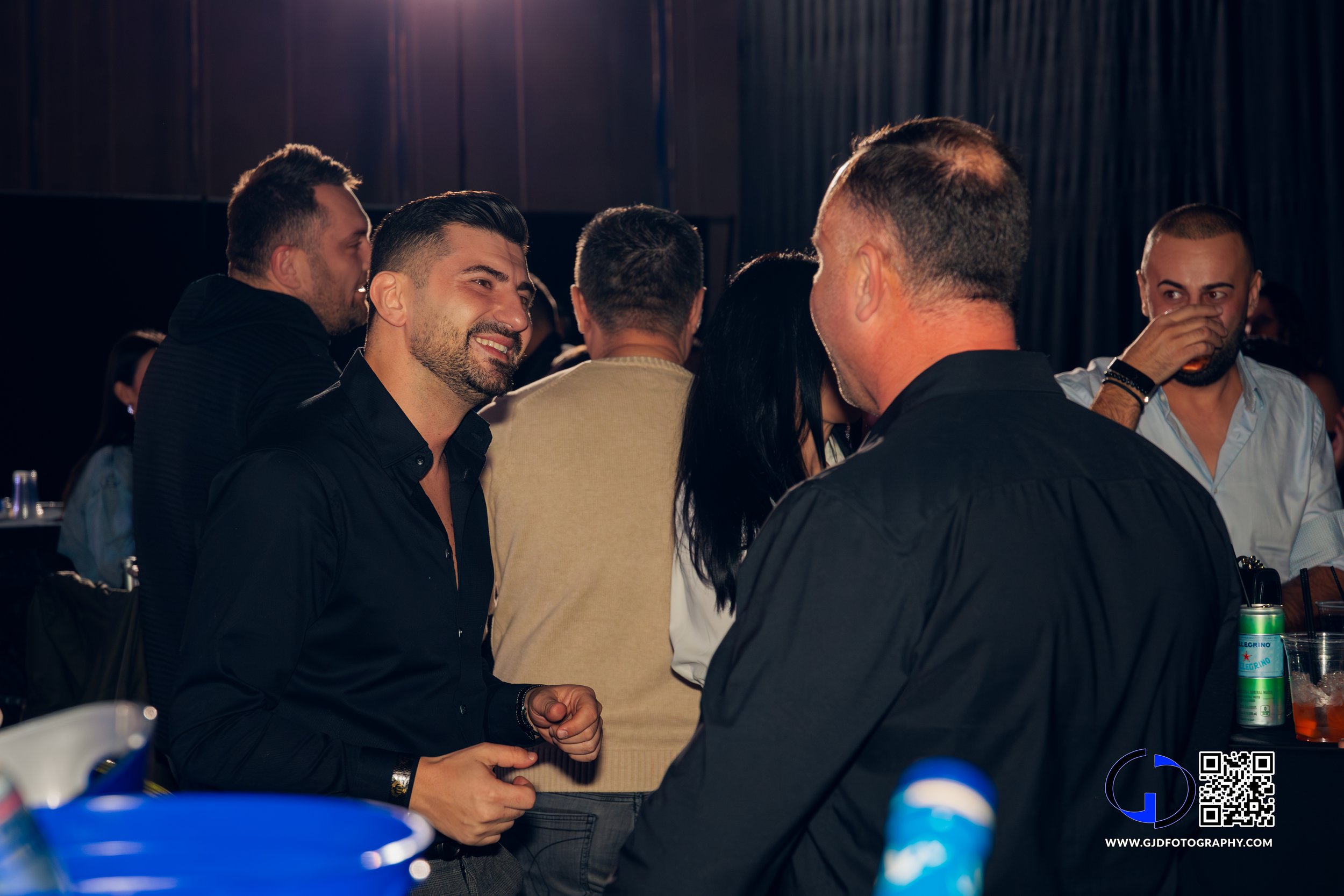 Group of people at a social gathering, talking and smiling in a dimly lit indoor venue.