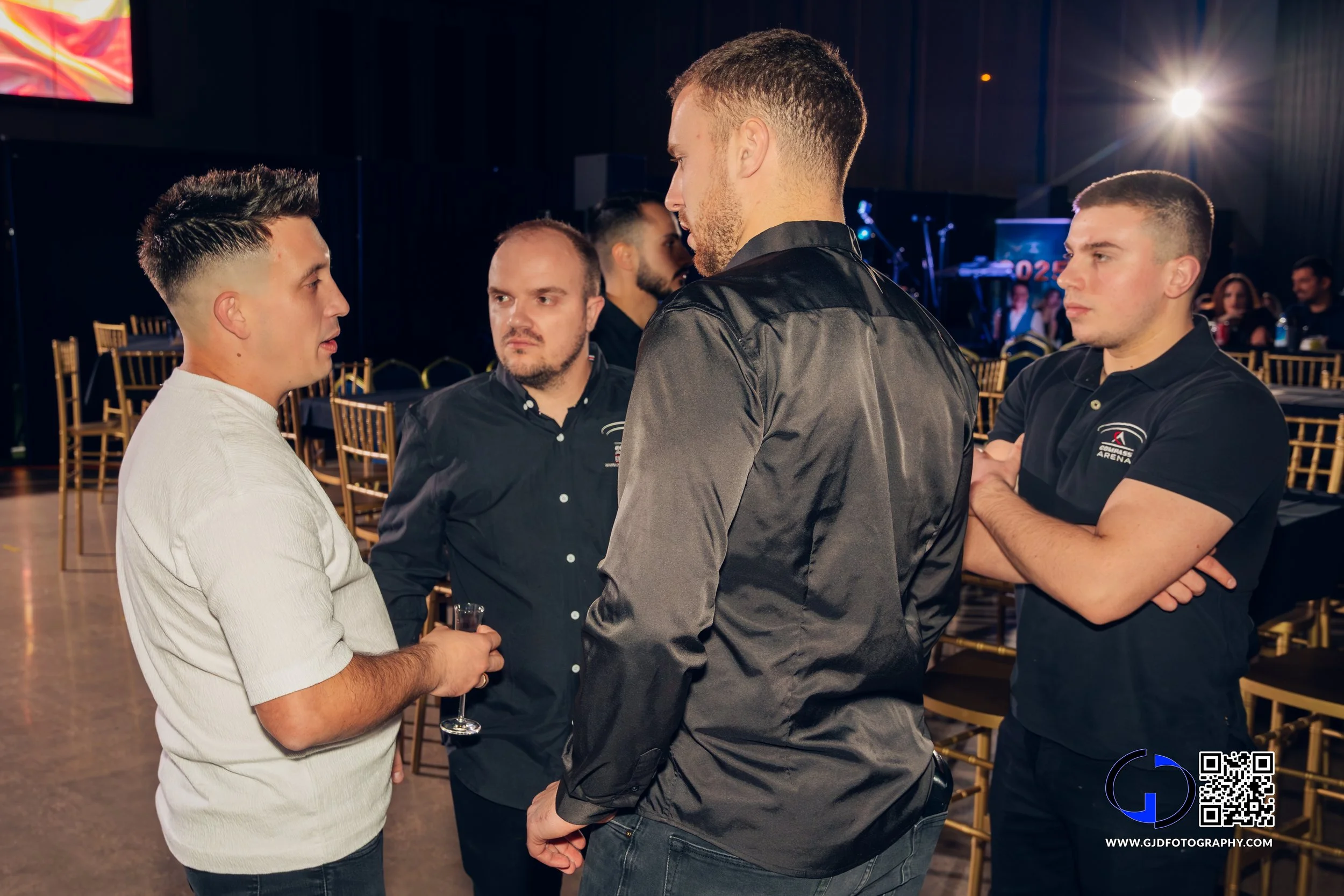 Four men engaged in a conversation at an indoor event, with empty chairs and a stage in the background.