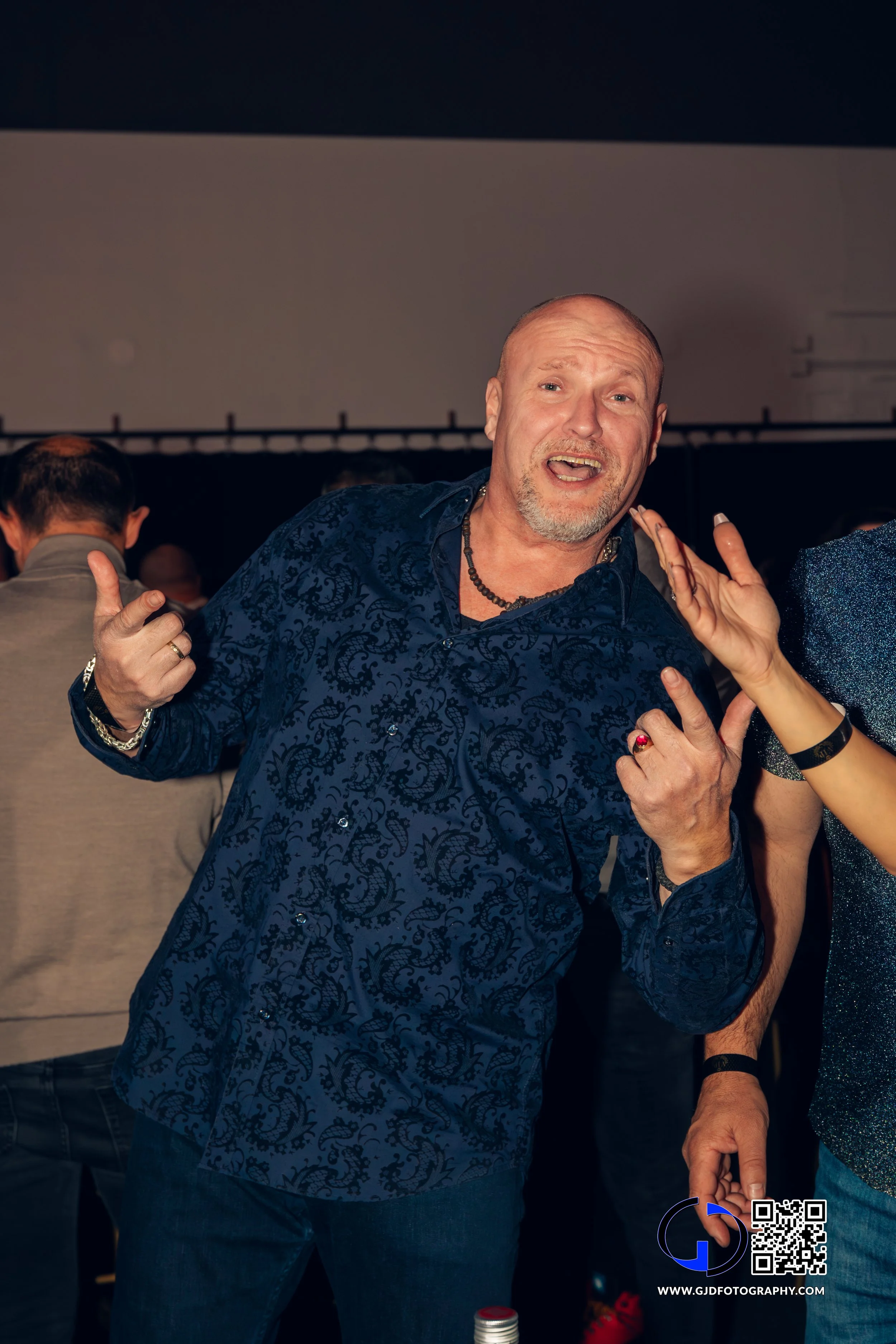 A man with a beard and short hair wearing a dark blue patterned shirt, making a fun gesture with his hands, at a social event or party.