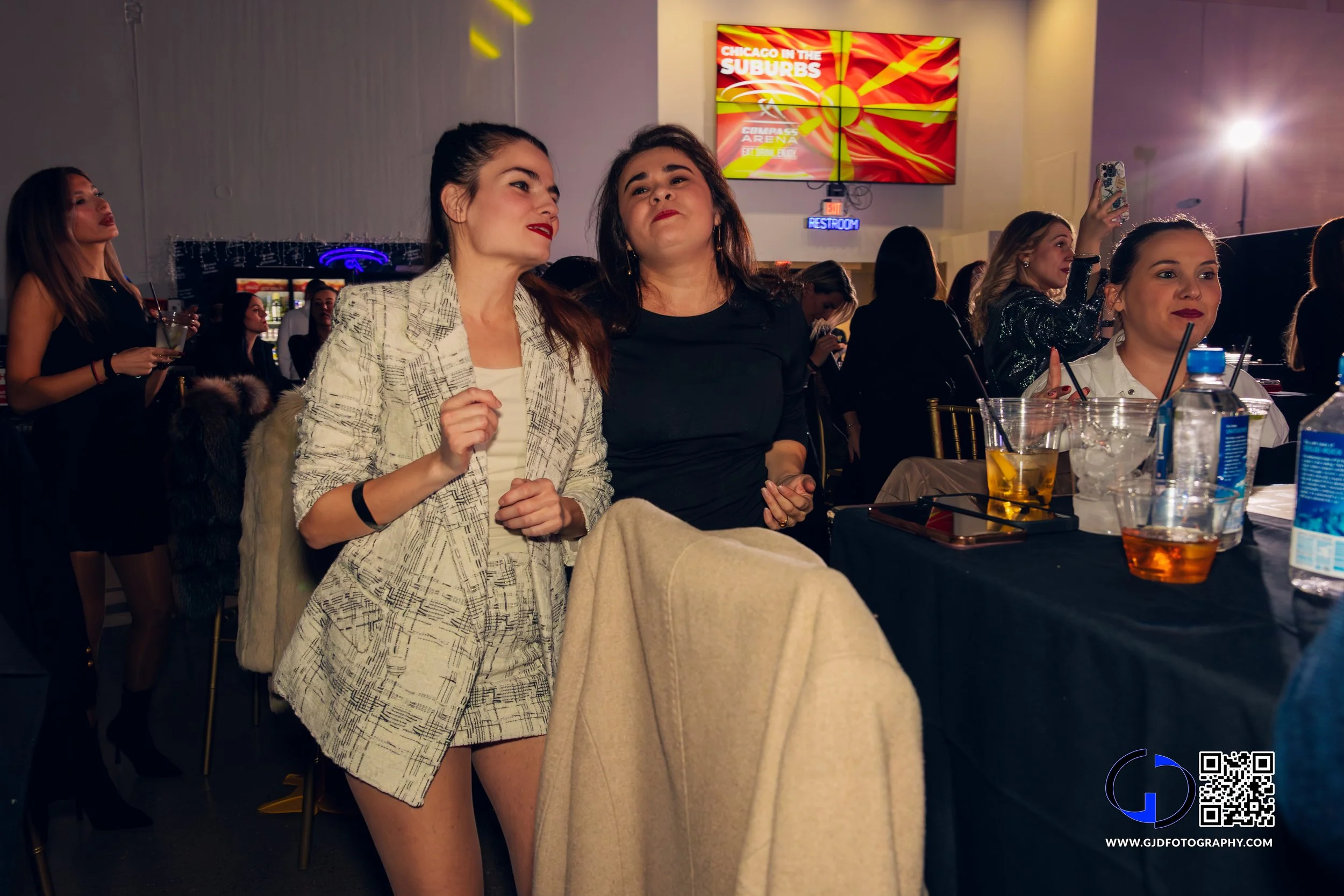Women at a lively indoor event, some dancing and others seated at a table with drinks, with colorful screens and neon signs in the background.