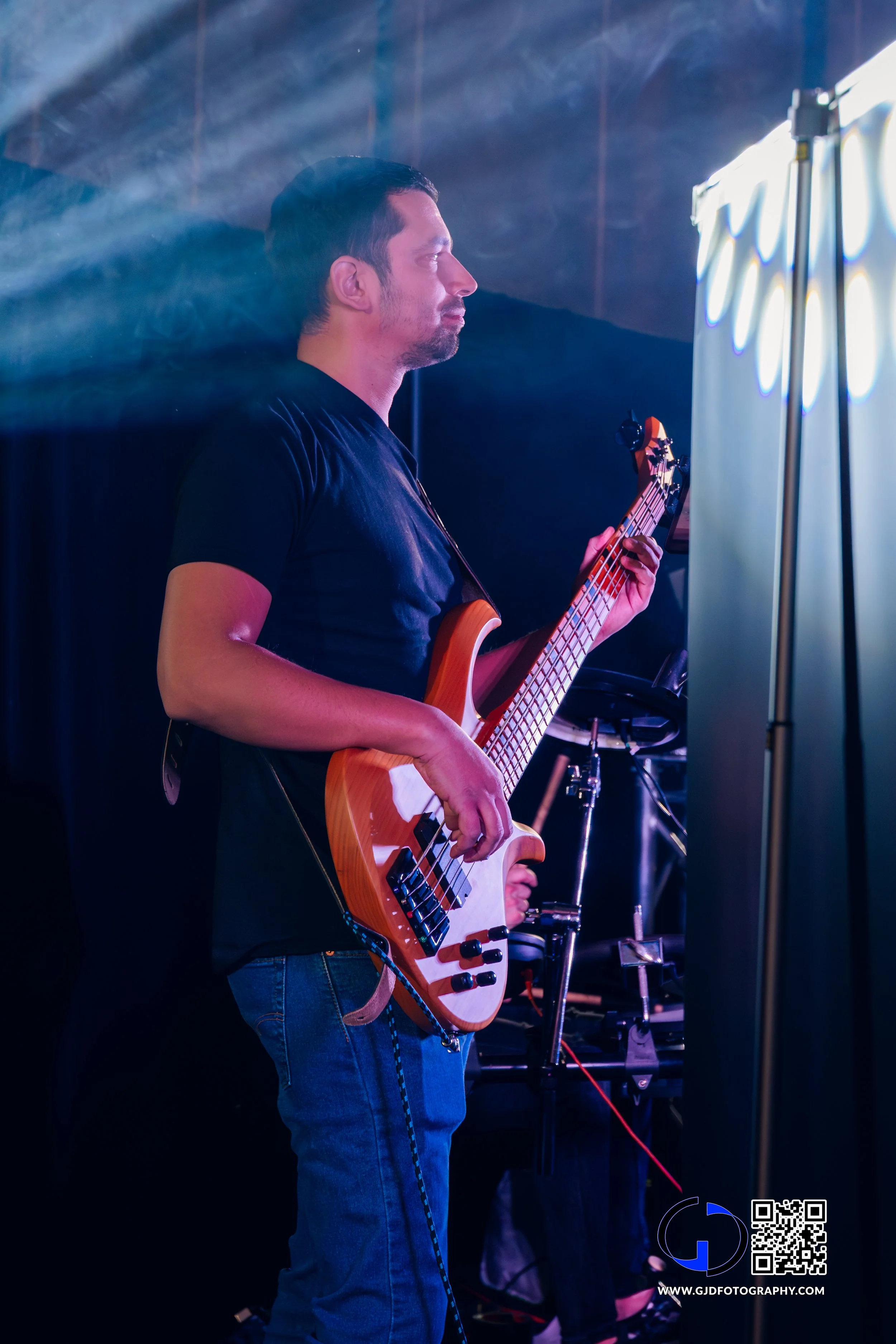 A man playing a bass guitar on stage, illuminated by stage lights, with a dark background and equipment nearby.