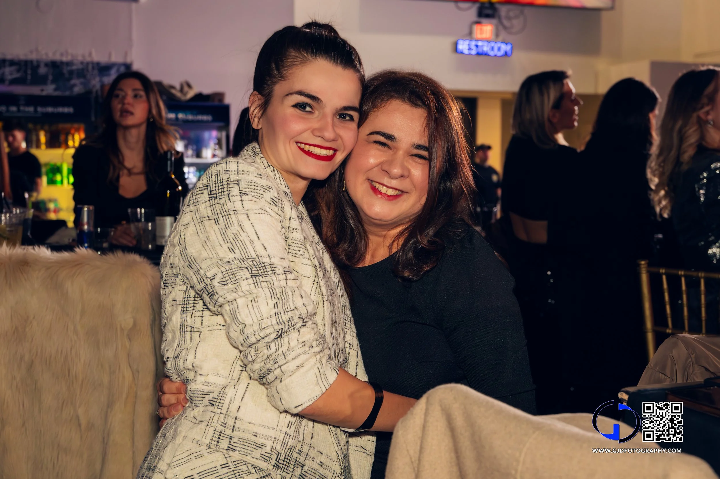 Two women smiling at a social gathering, seated close together with a bar in the background and other people standing and sitting.
