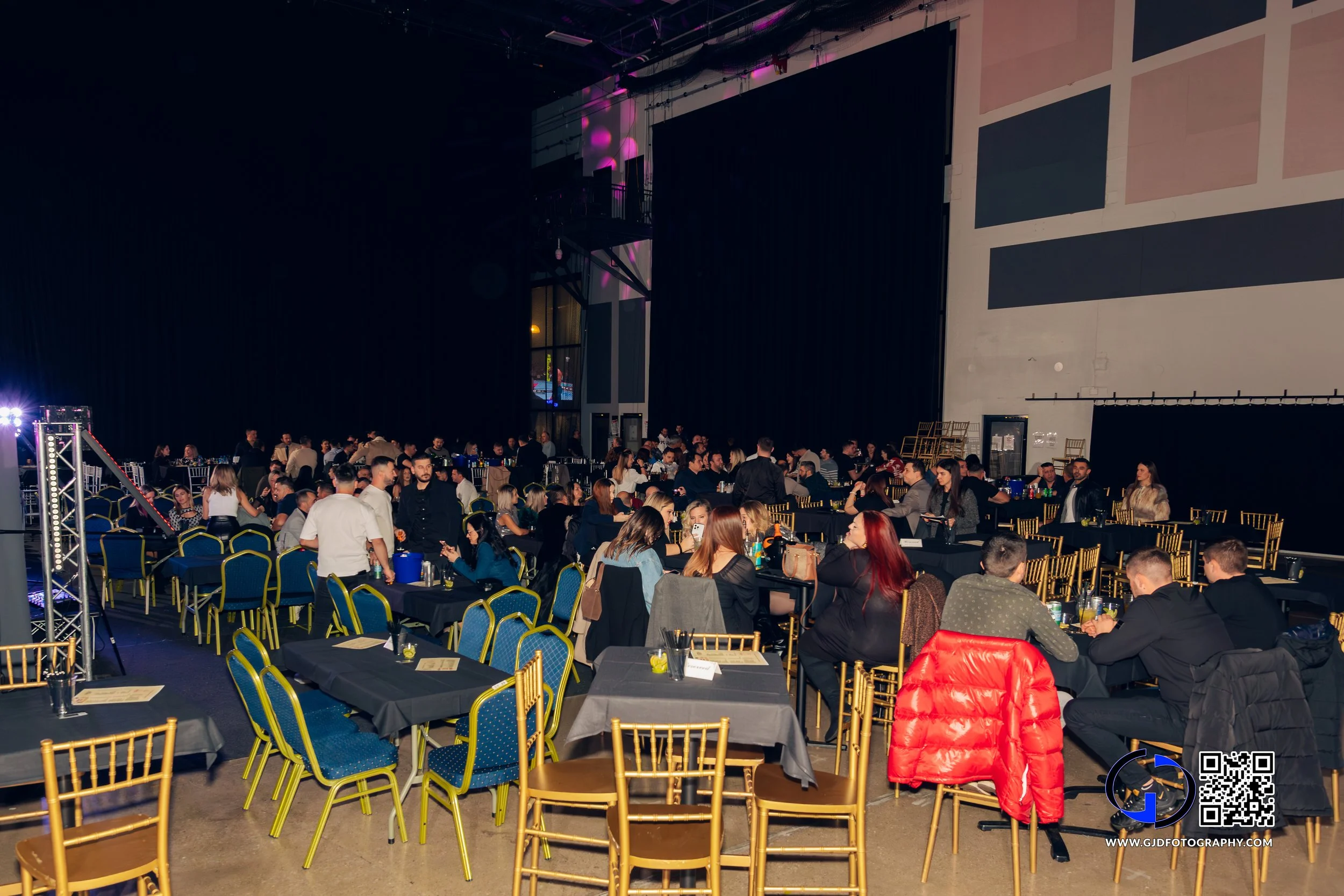 Large indoor event with many seated guests, black tablecloths, and gold and blue chairs in a spacious hall with black curtains and a high ceiling.