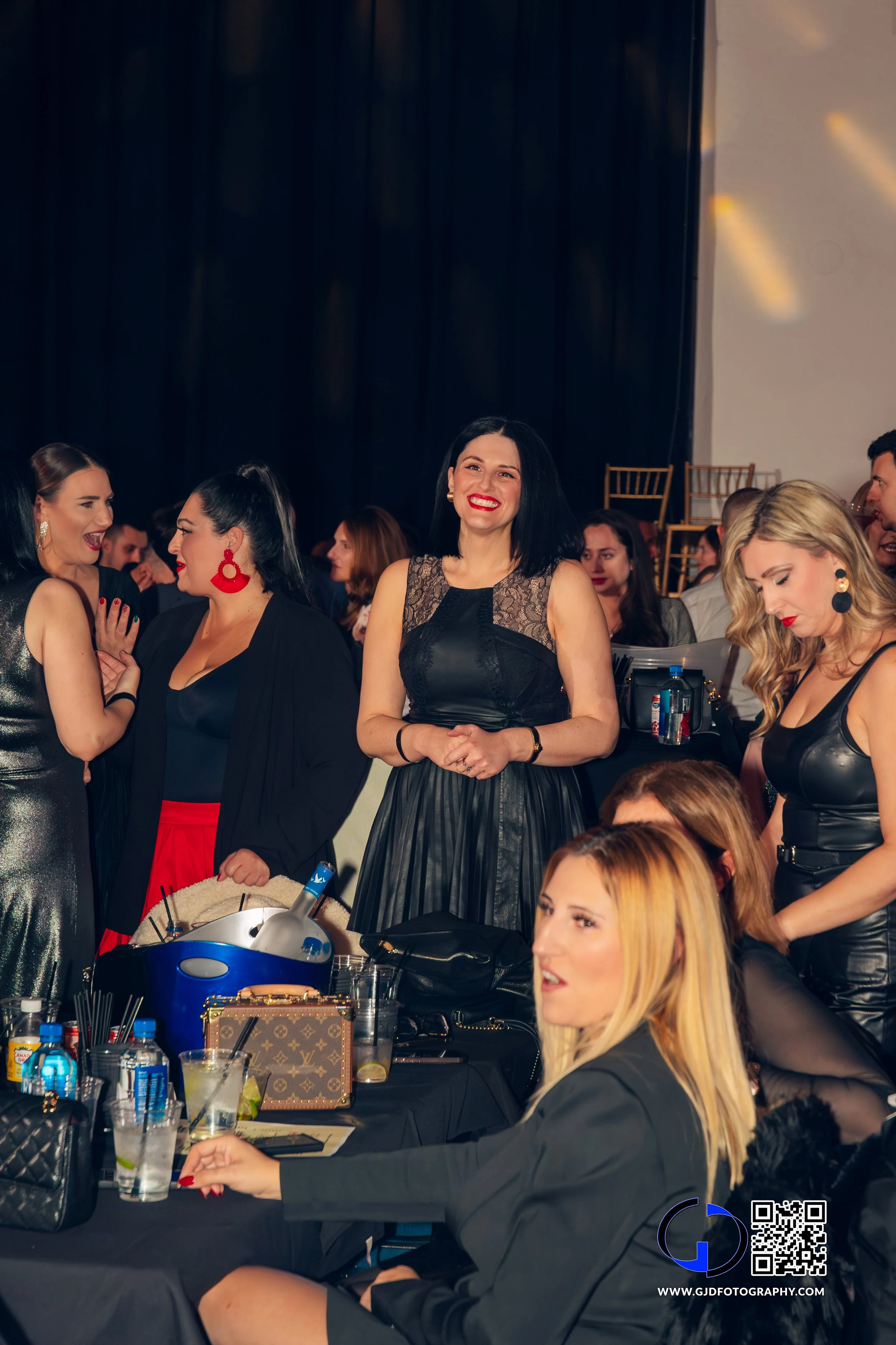 Group of women at a social event, some standing and some seated, dressed in fashionable attire, with drinks and accessories on the table.