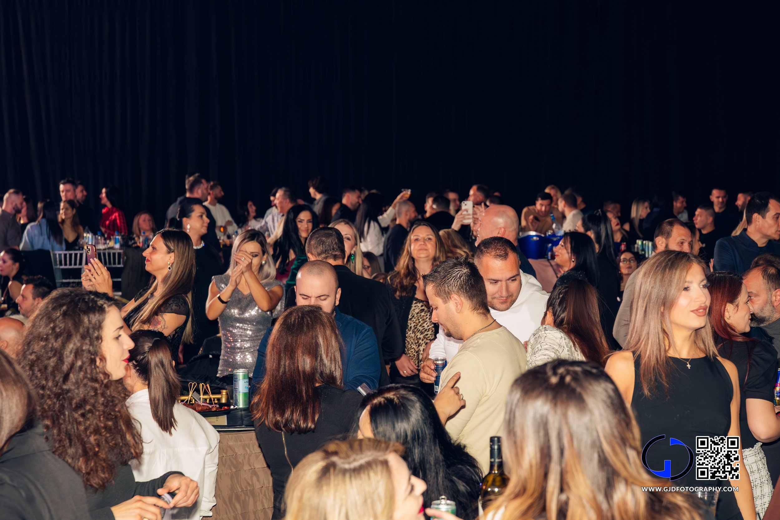 Crowd of people at an indoor event, some taking photos and mingling, with a dark background.