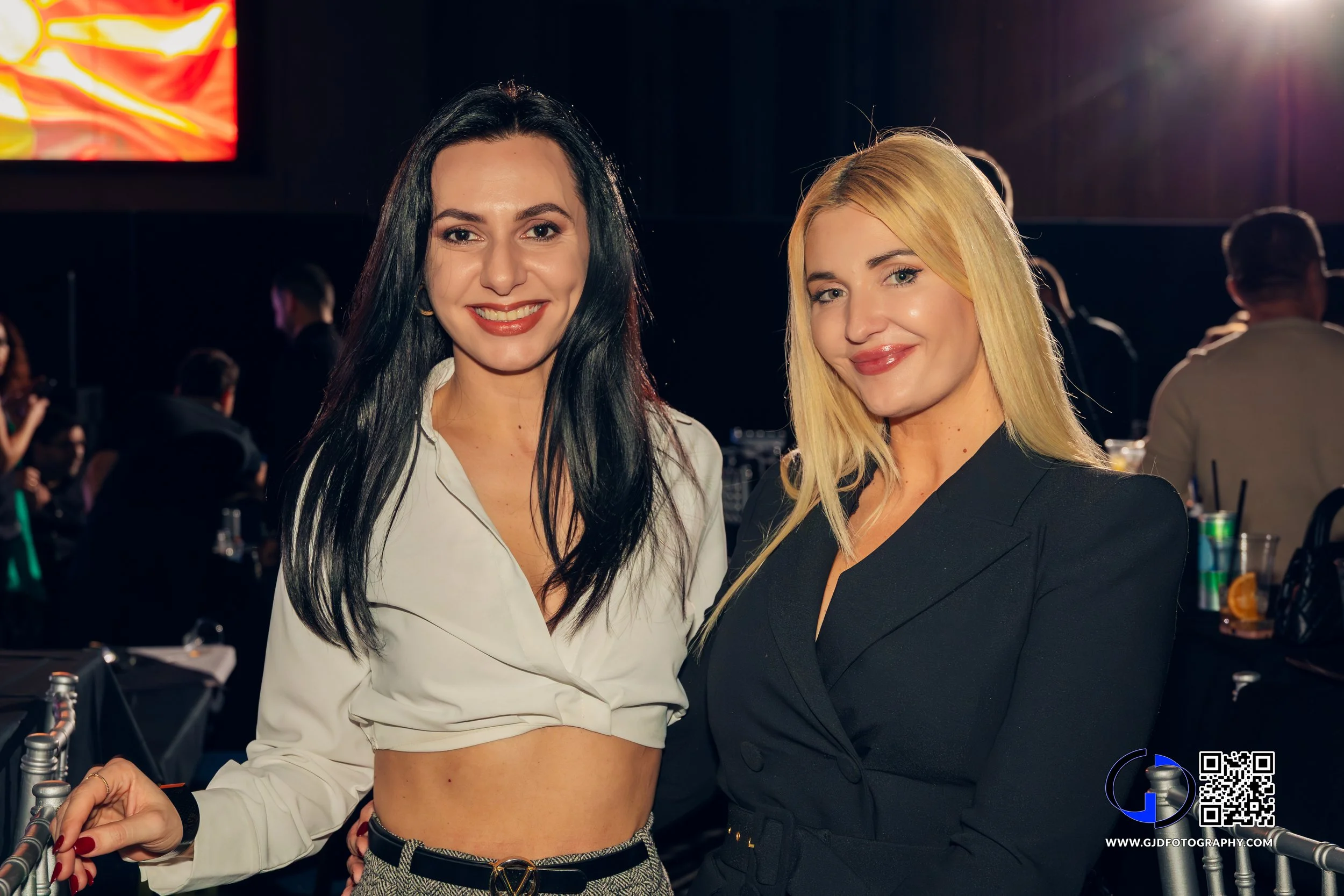 Two women smiling and standing close together at a social event or party, with a dark background and people mingling in the background.